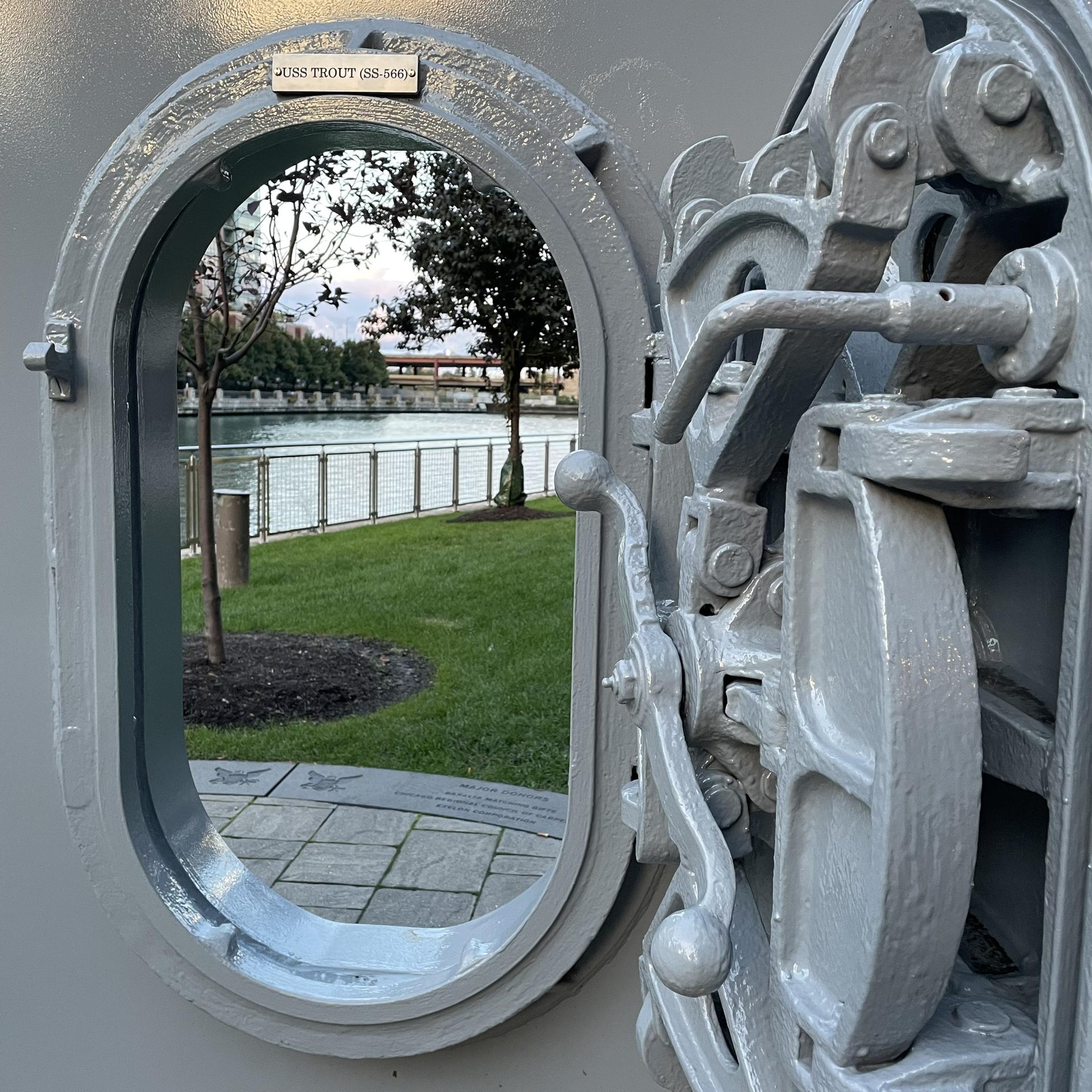 A gray porthole opens to a view of a calm river and green trees.