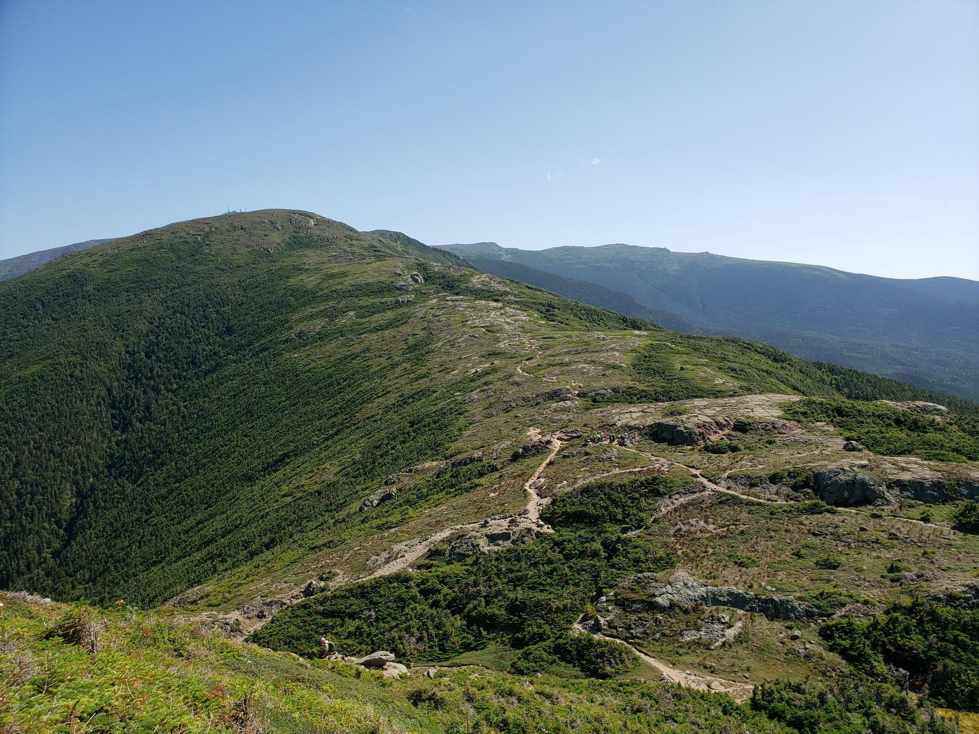 A winding trail ascends a green, rocky mountain under a clear blue sky.