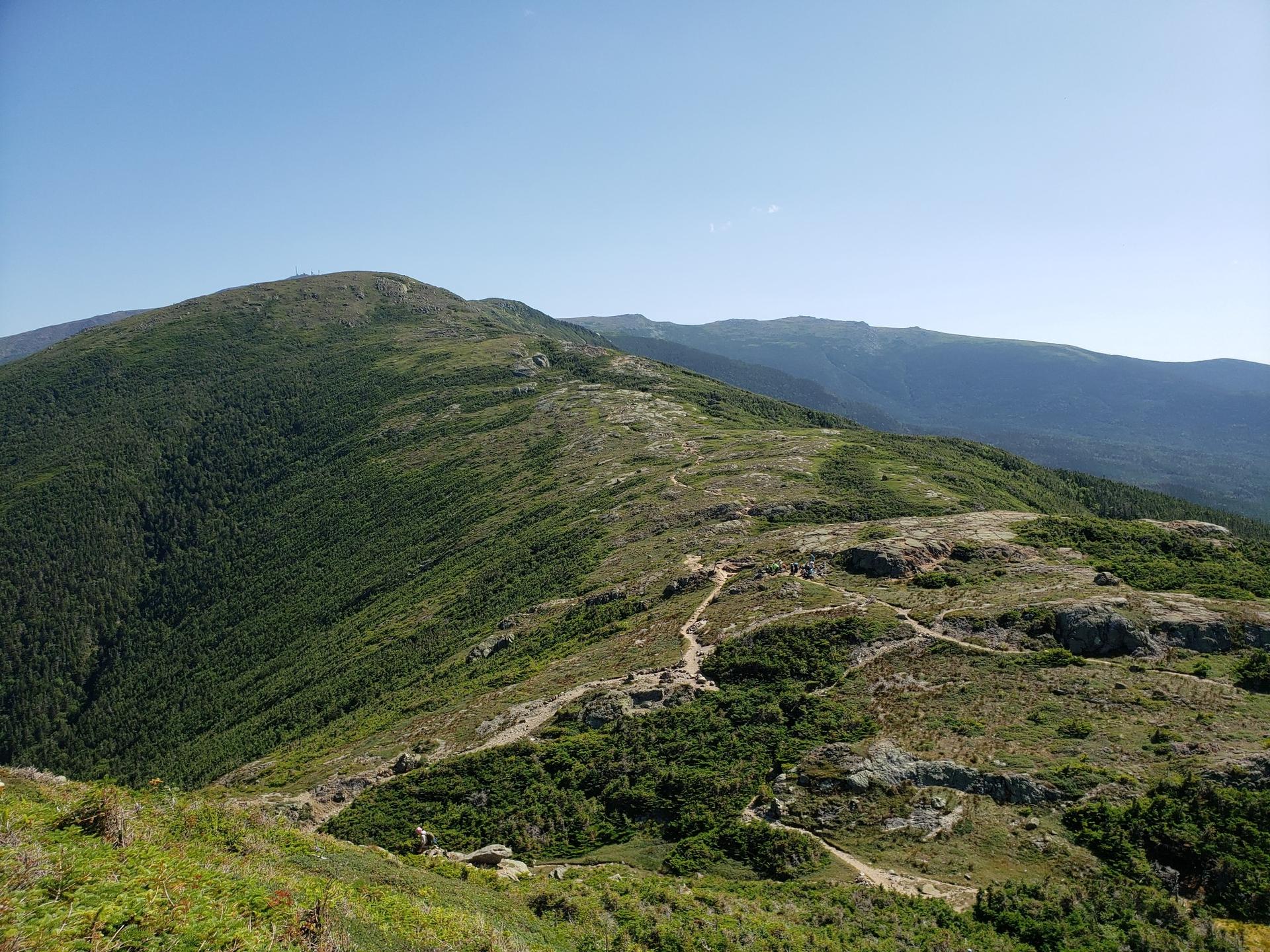A winding trail ascends a green, rocky mountain under a clear blue sky.