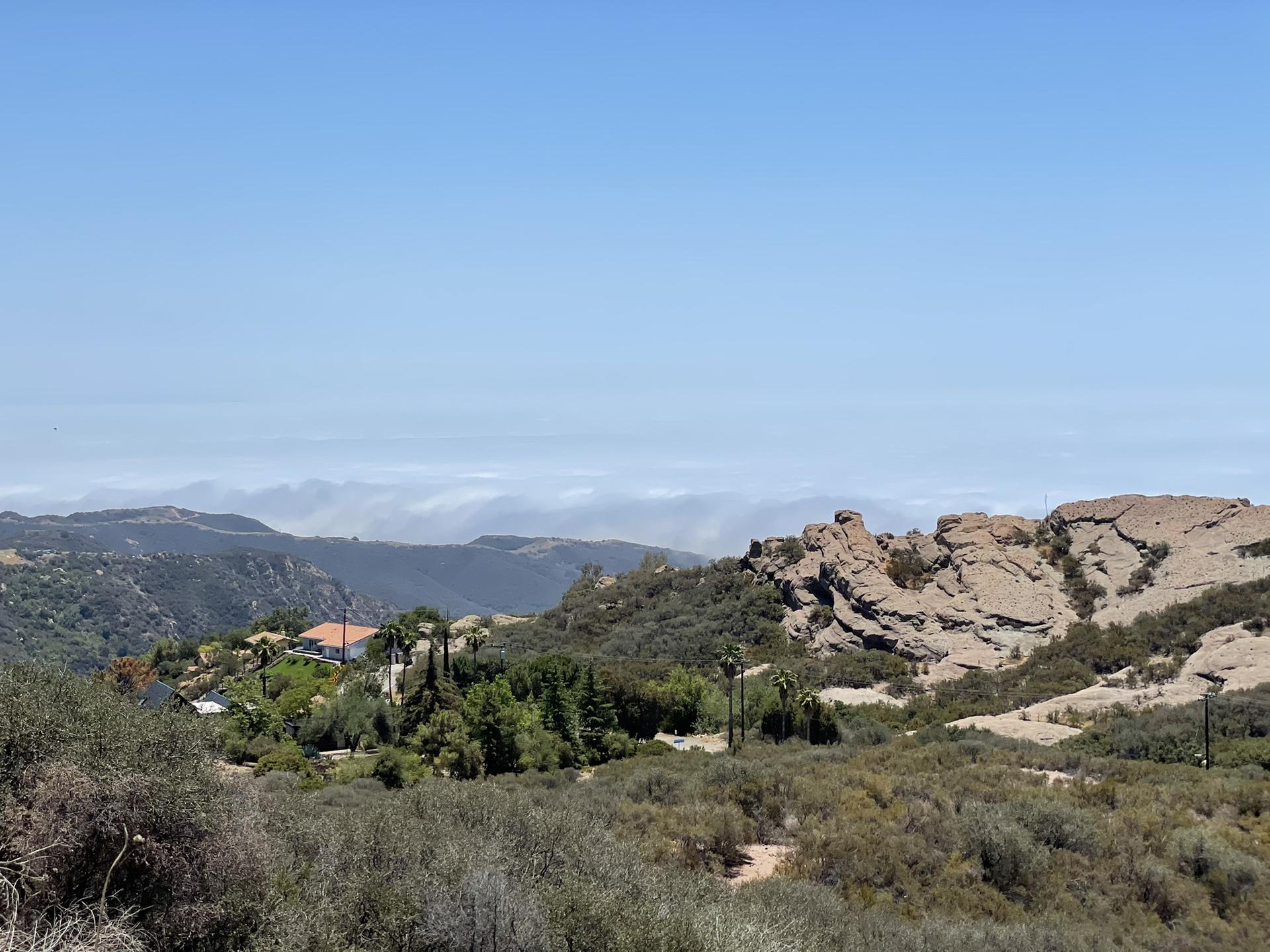 Bright mountain landscape with rocky outcrops, green trees, and distant hazy hills.