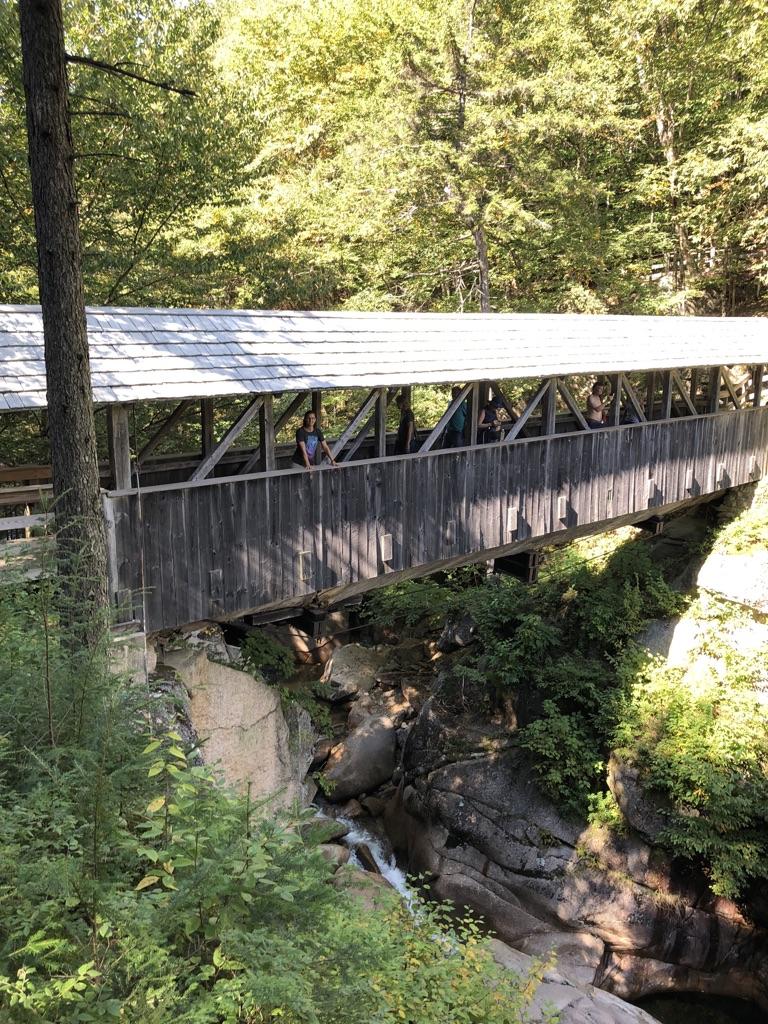 Wooden covered bridge over a rocky stream, people visible inside.