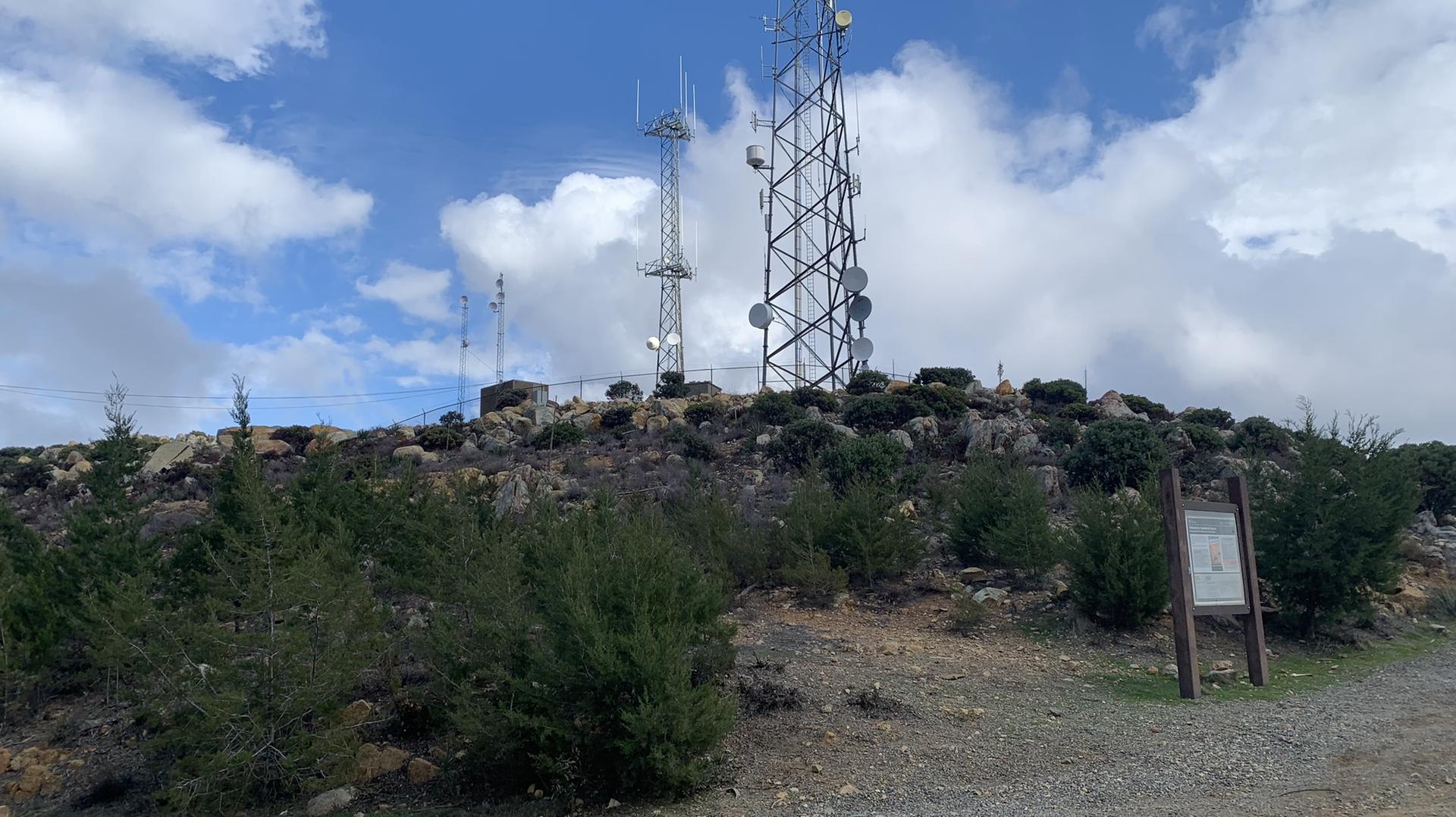 Communication towers on a rocky, scrubby hill under a bright, cloudy sky.