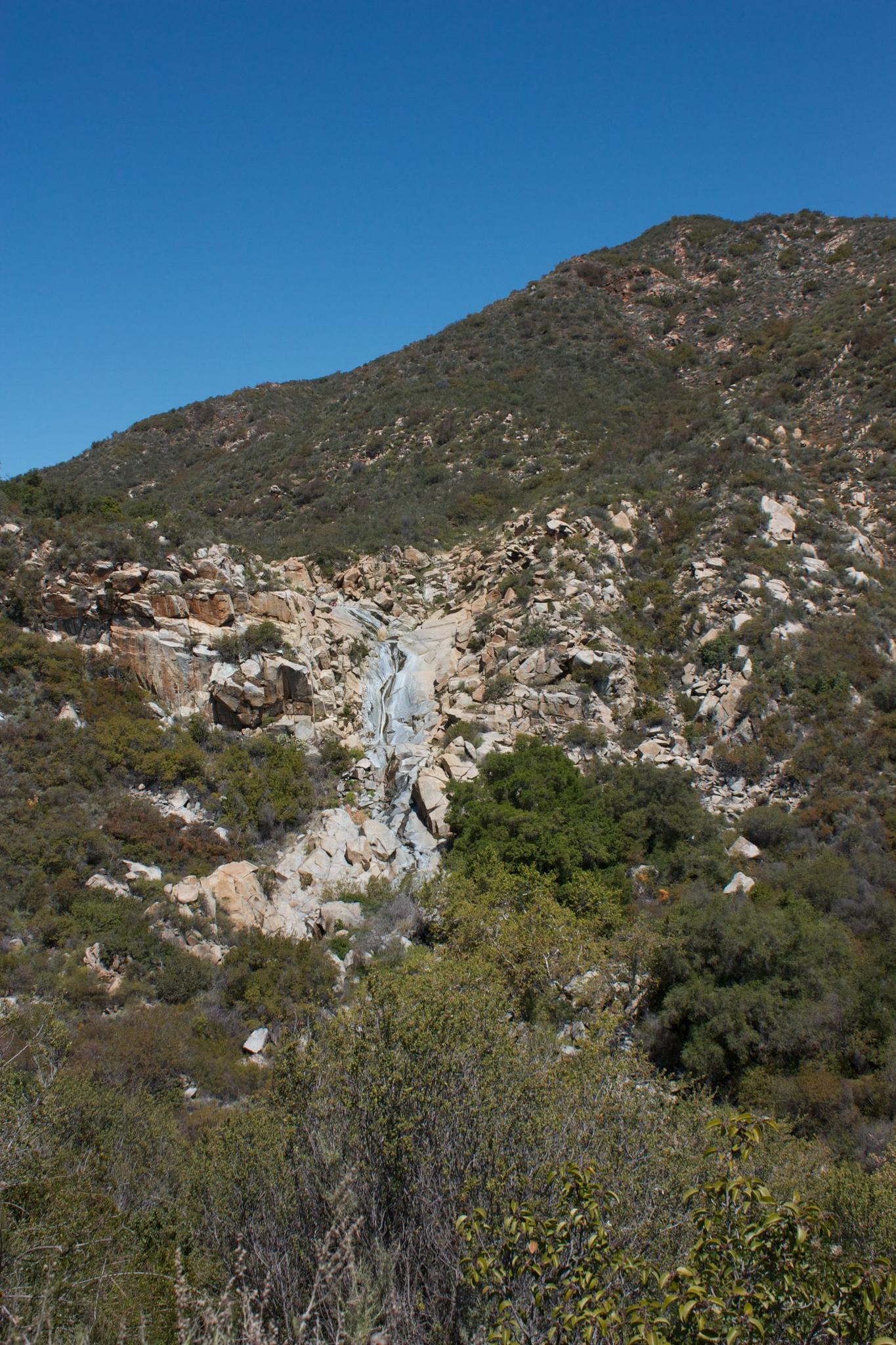 Small waterfall cascading down a rocky, brush-covered mountainside under clear sky.