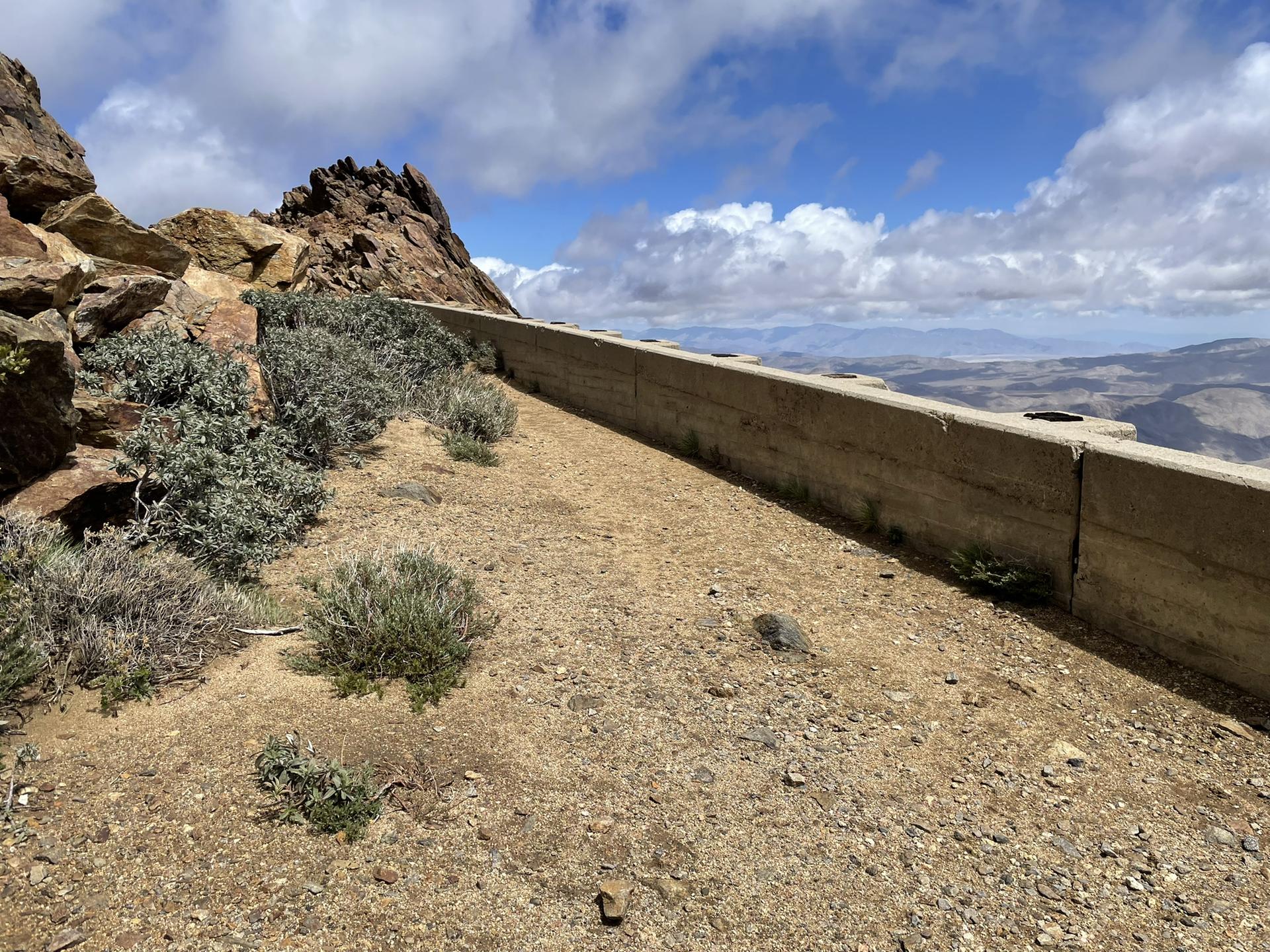 Dirt path next to a concrete barrier on a mountain ridge with distant views.