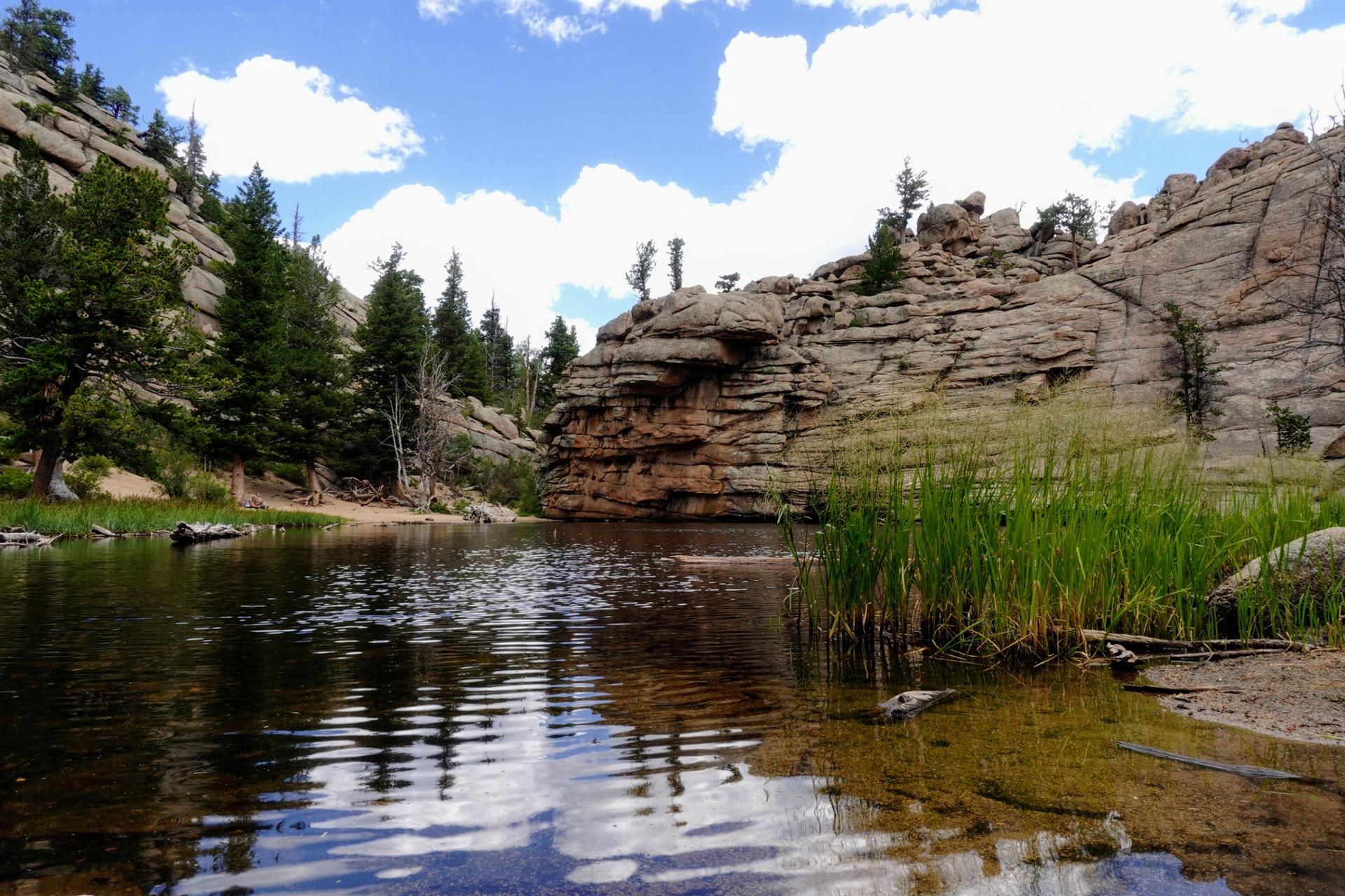A serene lake reflecting the sky, bordered by rocky cliffs and tall grass.