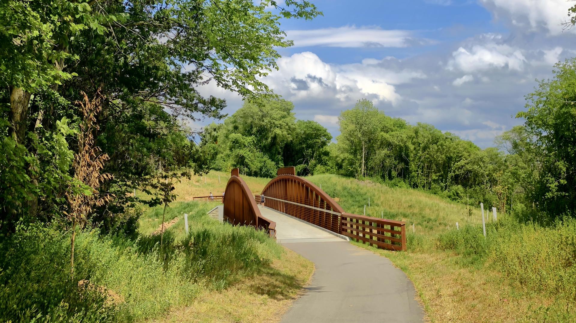 Paved path leading to a wooden arched bridge over green grass, cloudy sky.