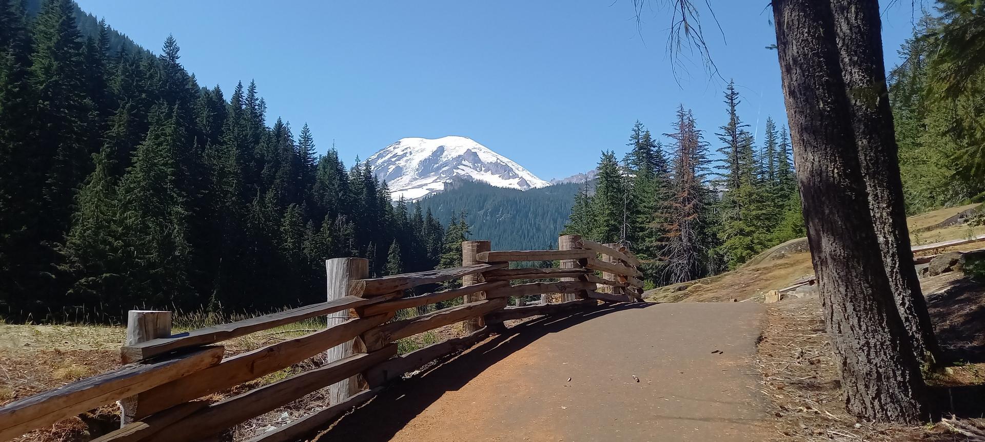 A paved path with a wooden fence leads towards a distant snow-capped mountain.
