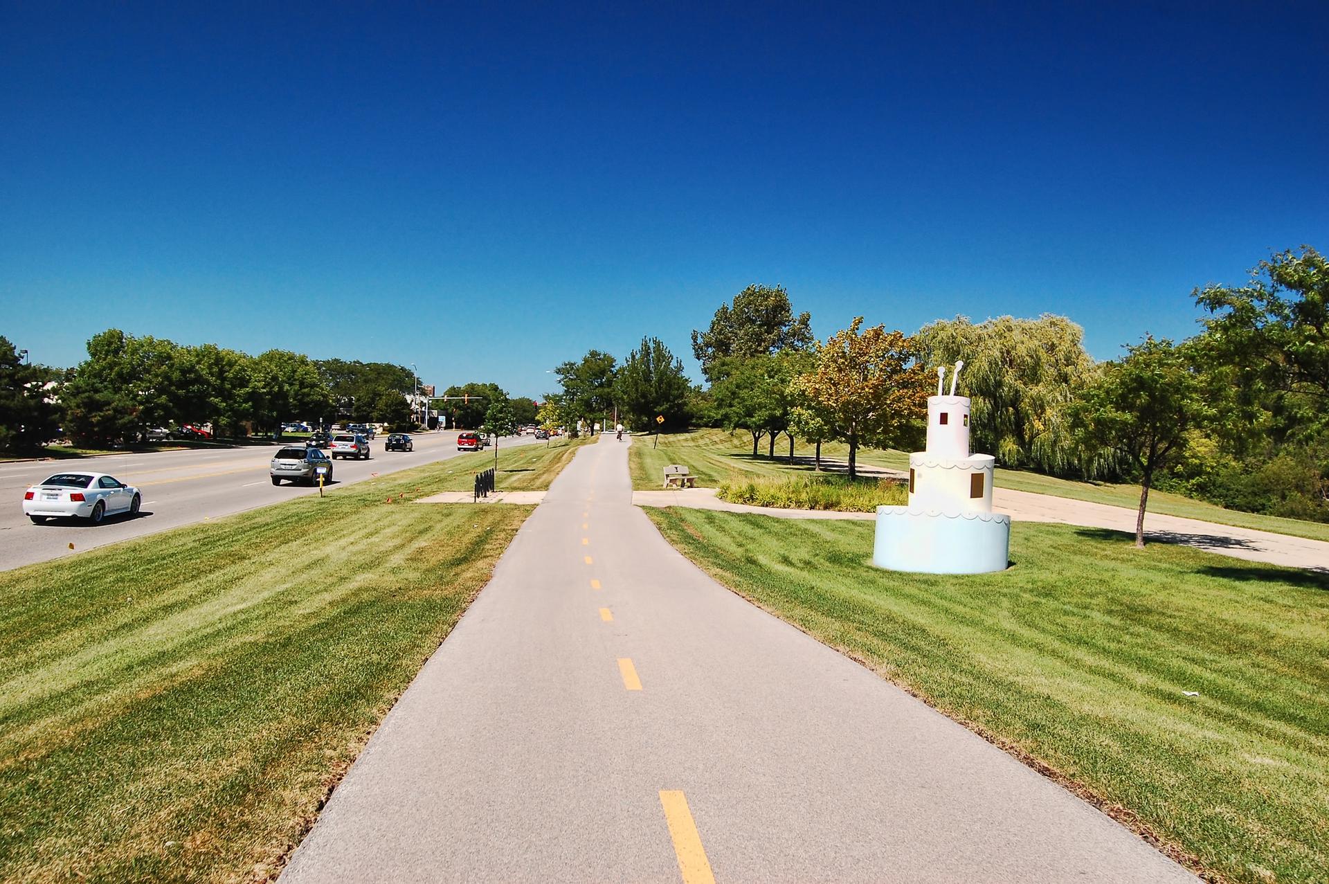 Paved path, grassy verges, road with cars, and a white sculpture on a sunny day.