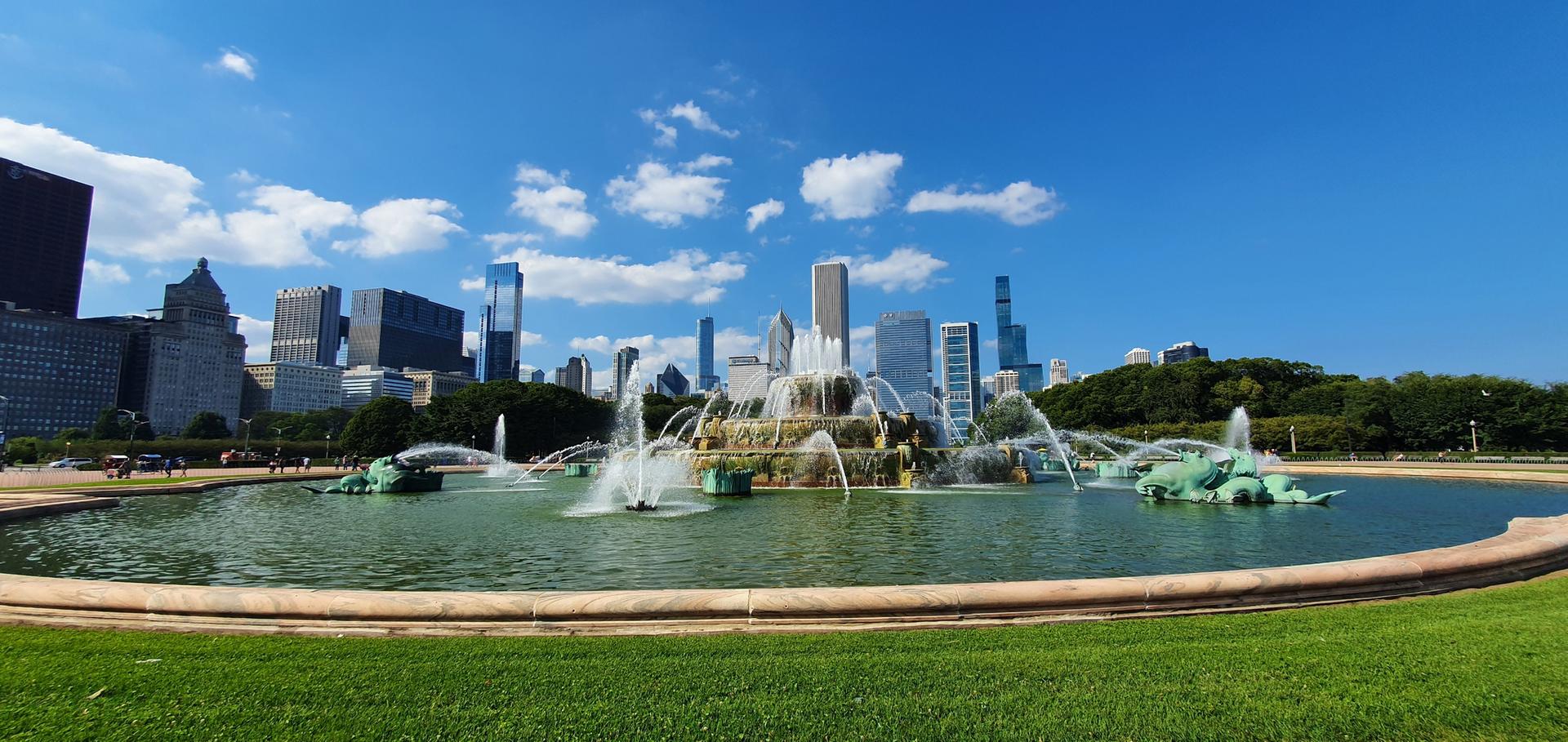 Large fountain spraying water, with a city skyline and blue sky in the background.