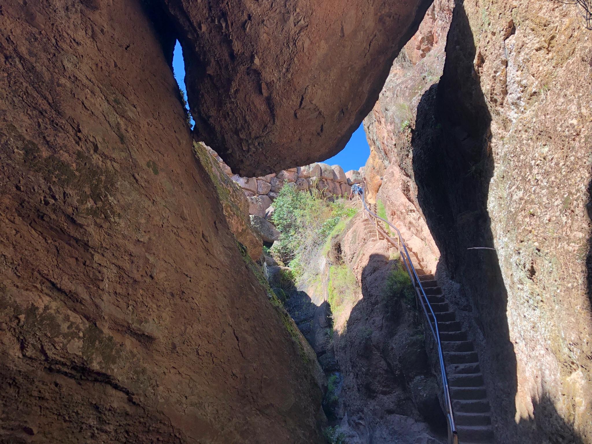 Narrow rock canyon with stairs leading up, sunlight filtering through a rock arch.