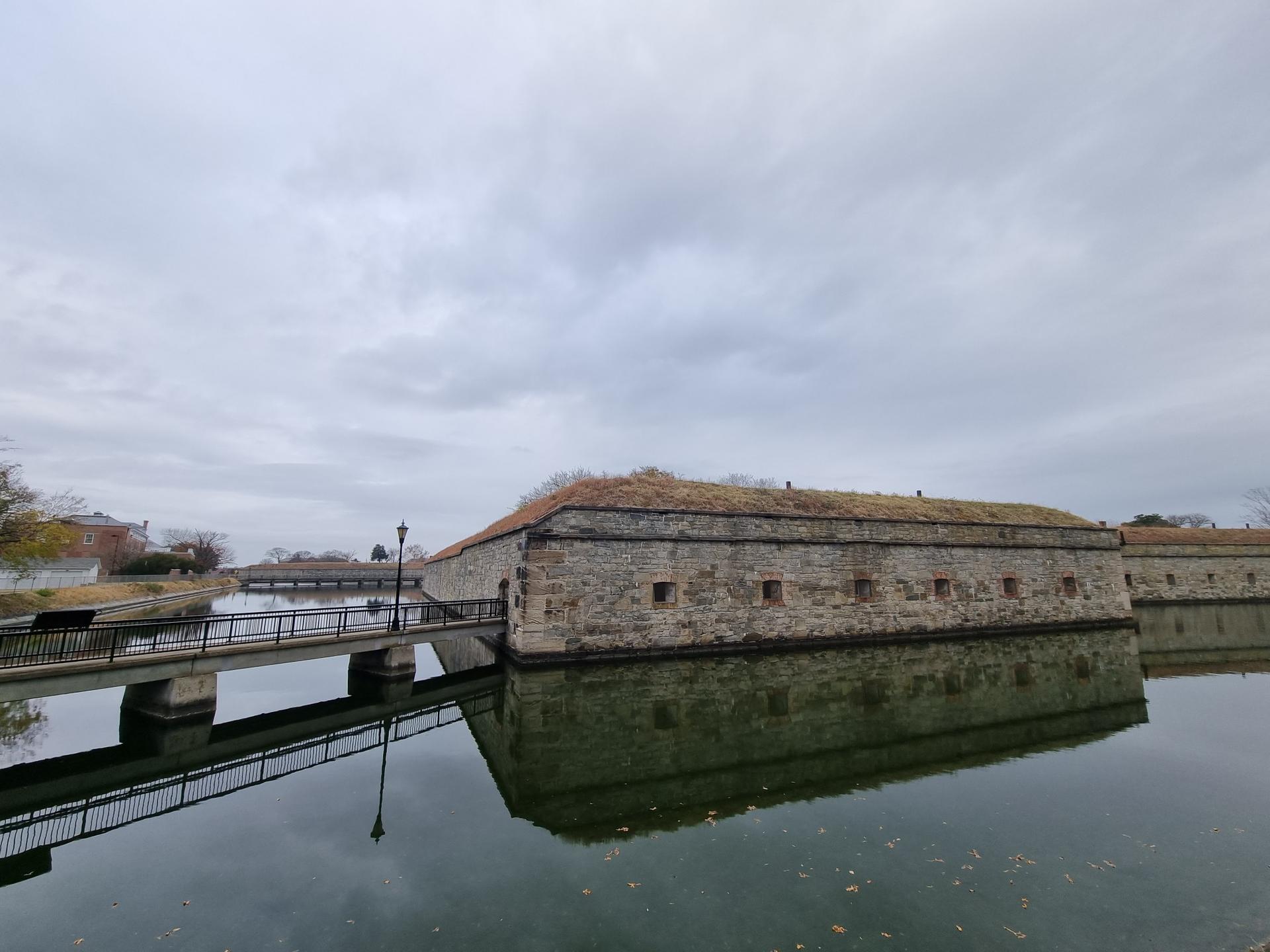 Stone structure with grass roof reflected in calm water under an overcast sky.