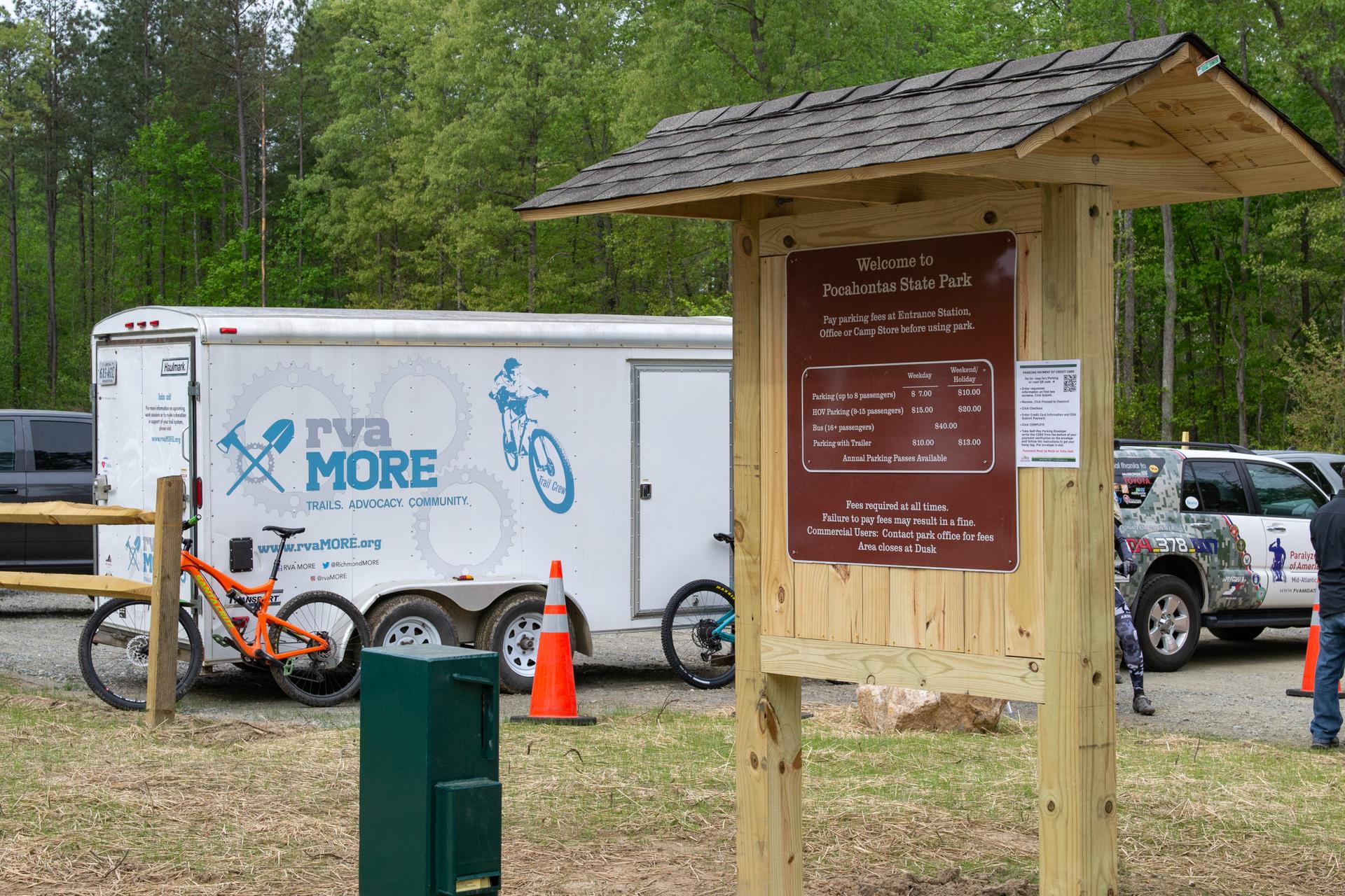 Wooden sign, trailer, and mountain bike in a wooded area on a bright day.