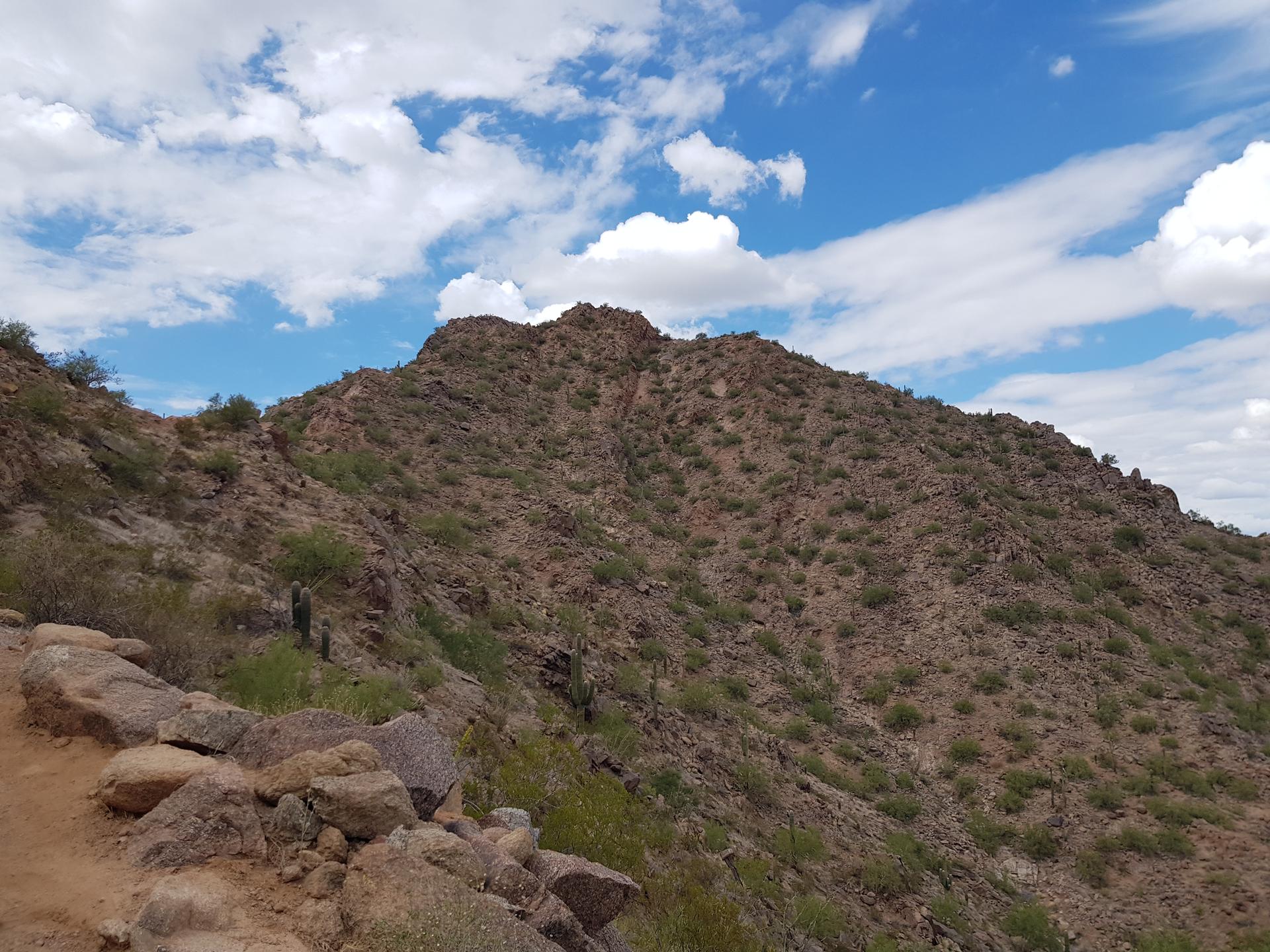 Steep, rocky desert mountain covered in sparse green vegetation under a cloudy sky.