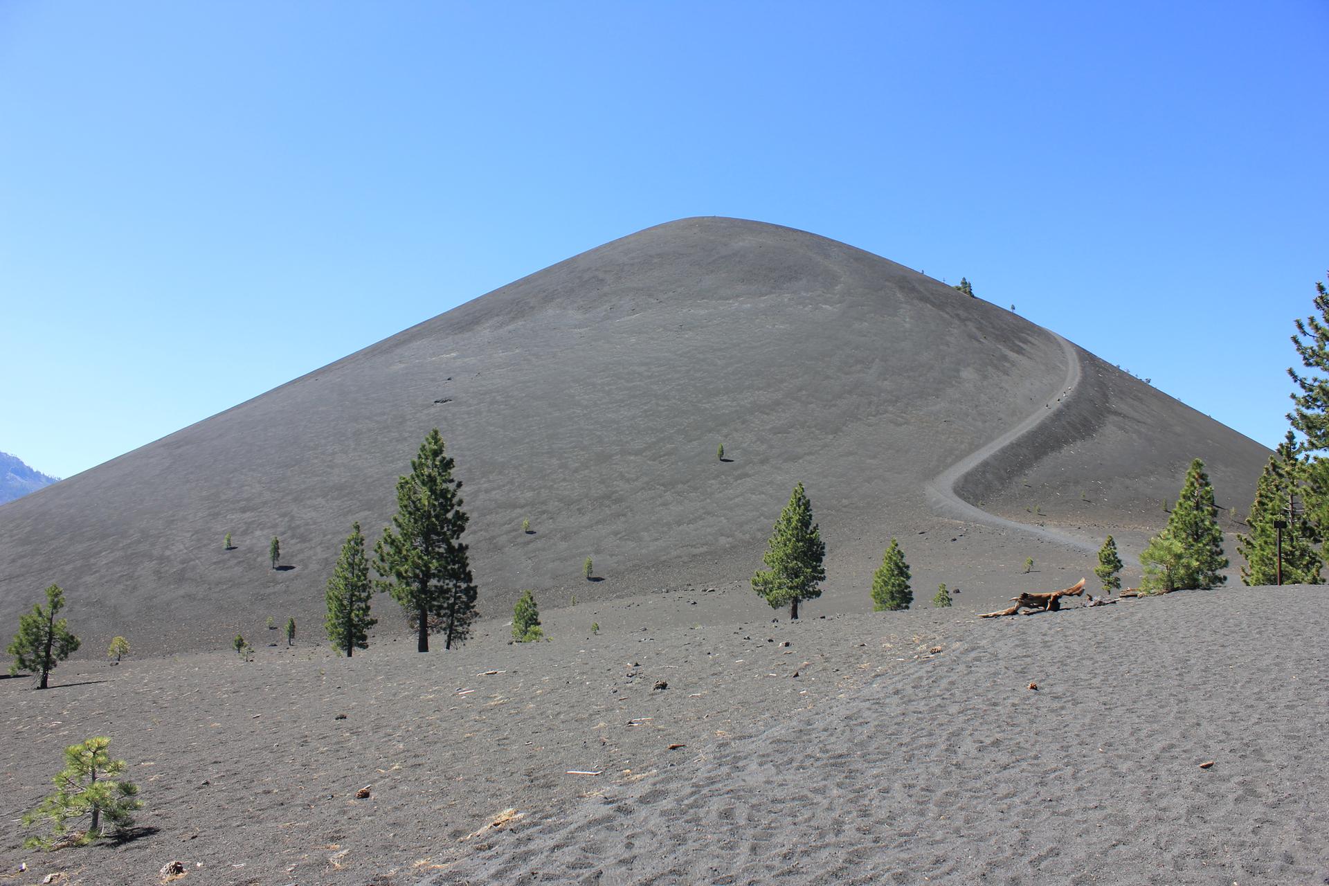 A large, dark cinder cone with a winding trail under a clear blue sky.