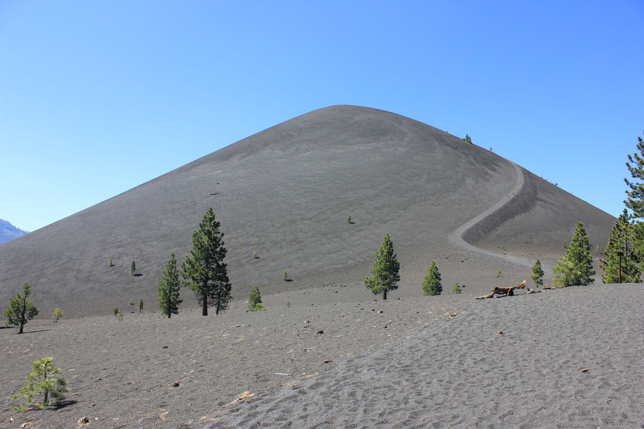 A large, dark cinder cone with a winding trail under a clear blue sky. - Cinder Cone Trail