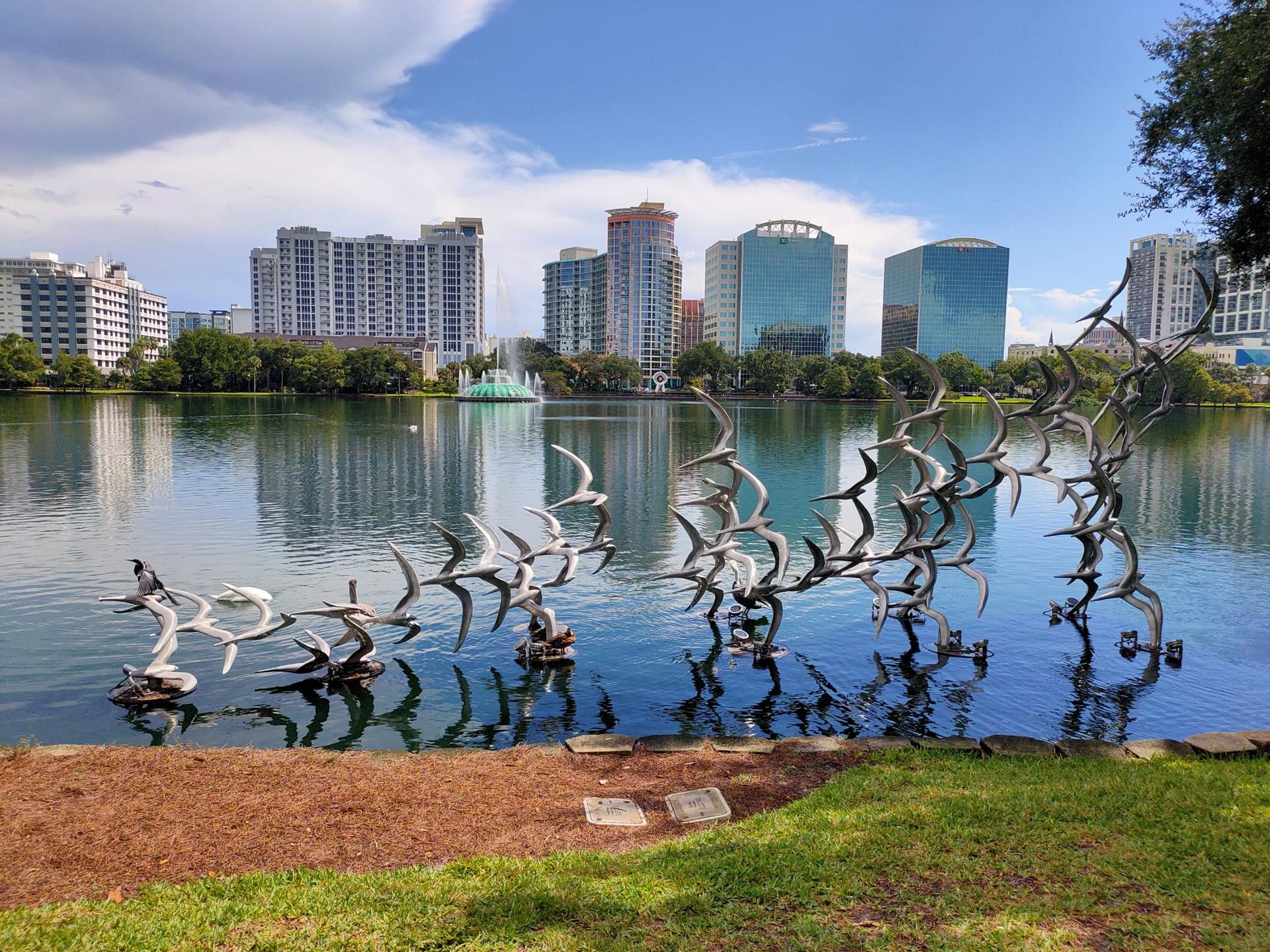 Metal bird sculptures in a lake with city buildings under a bright sky.