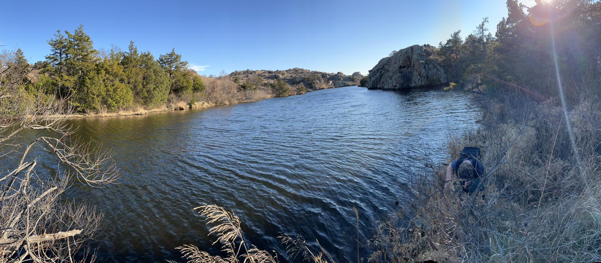 Wide river flows between rocky, wooded shores under bright sun. A person crouches.