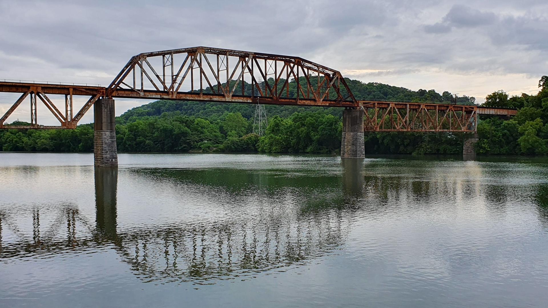 A rusty metal truss bridge crosses a calm river with forested hills.