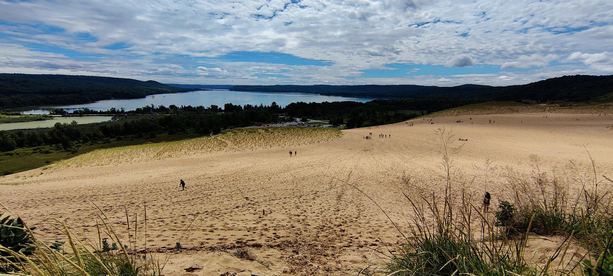 Expansive sand dunes with people walking, overlooking a large lake under a cloudy sky.