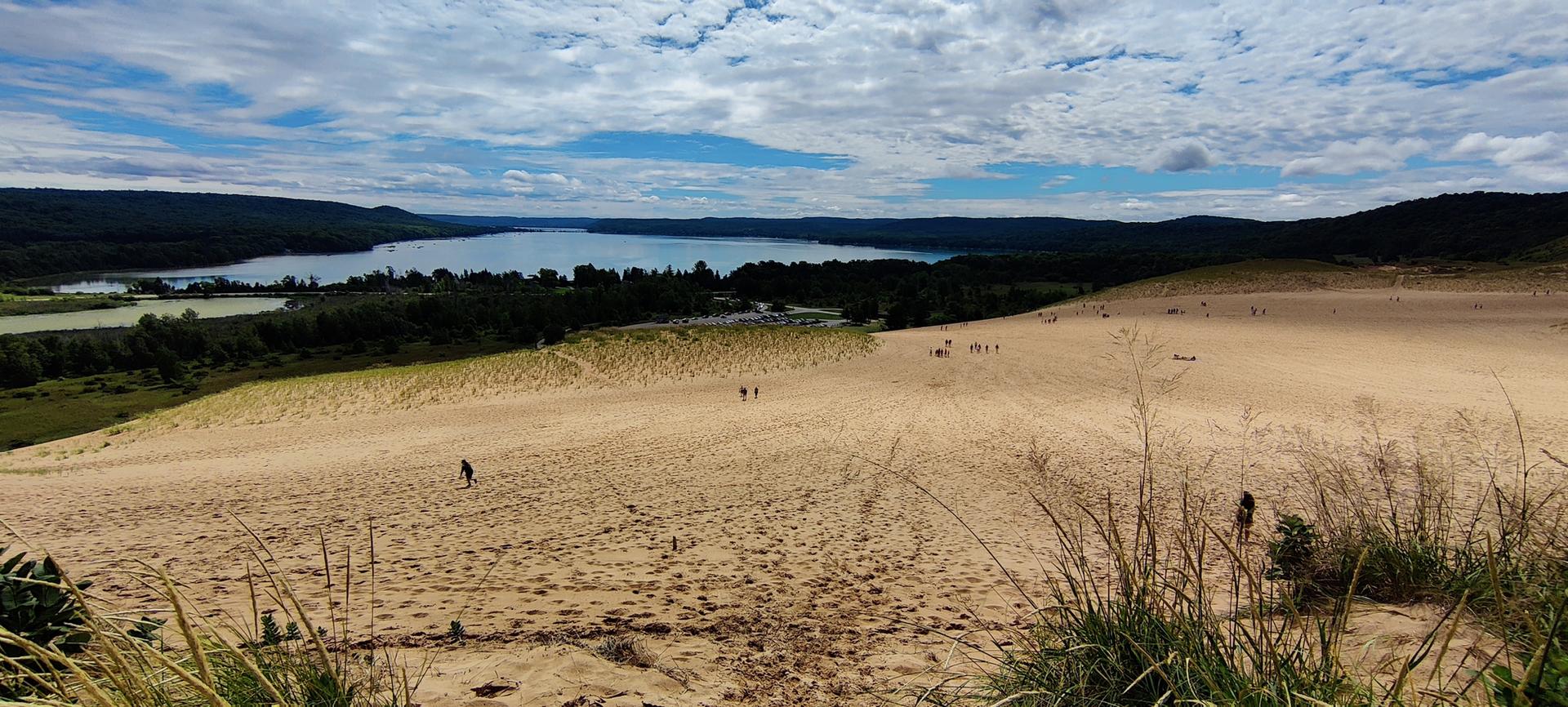 Expansive sand dunes with people walking, overlooking a large lake under a cloudy sky.