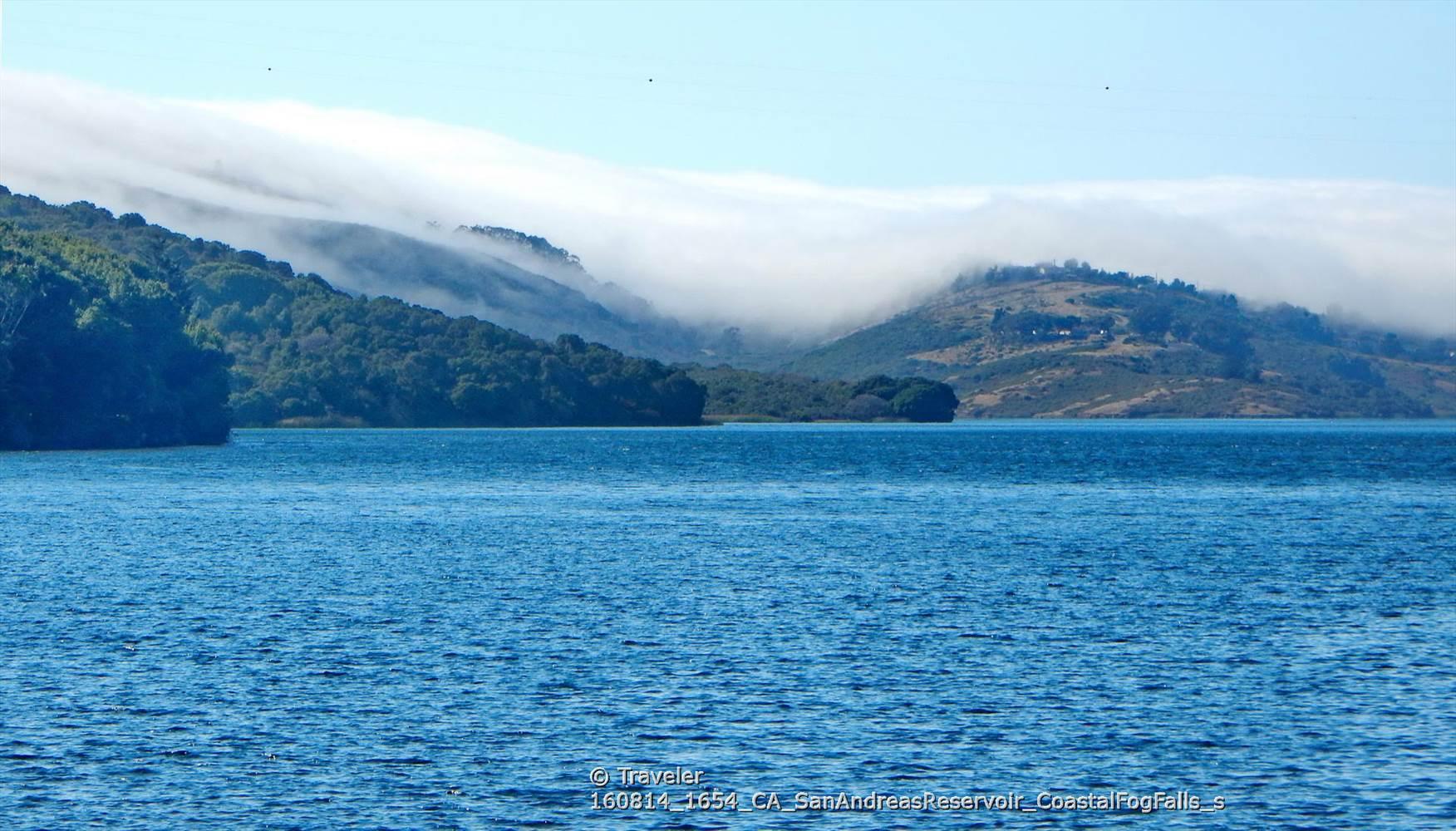 Blue water with forested hills partially covered by low-lying mist under a bright sky.
