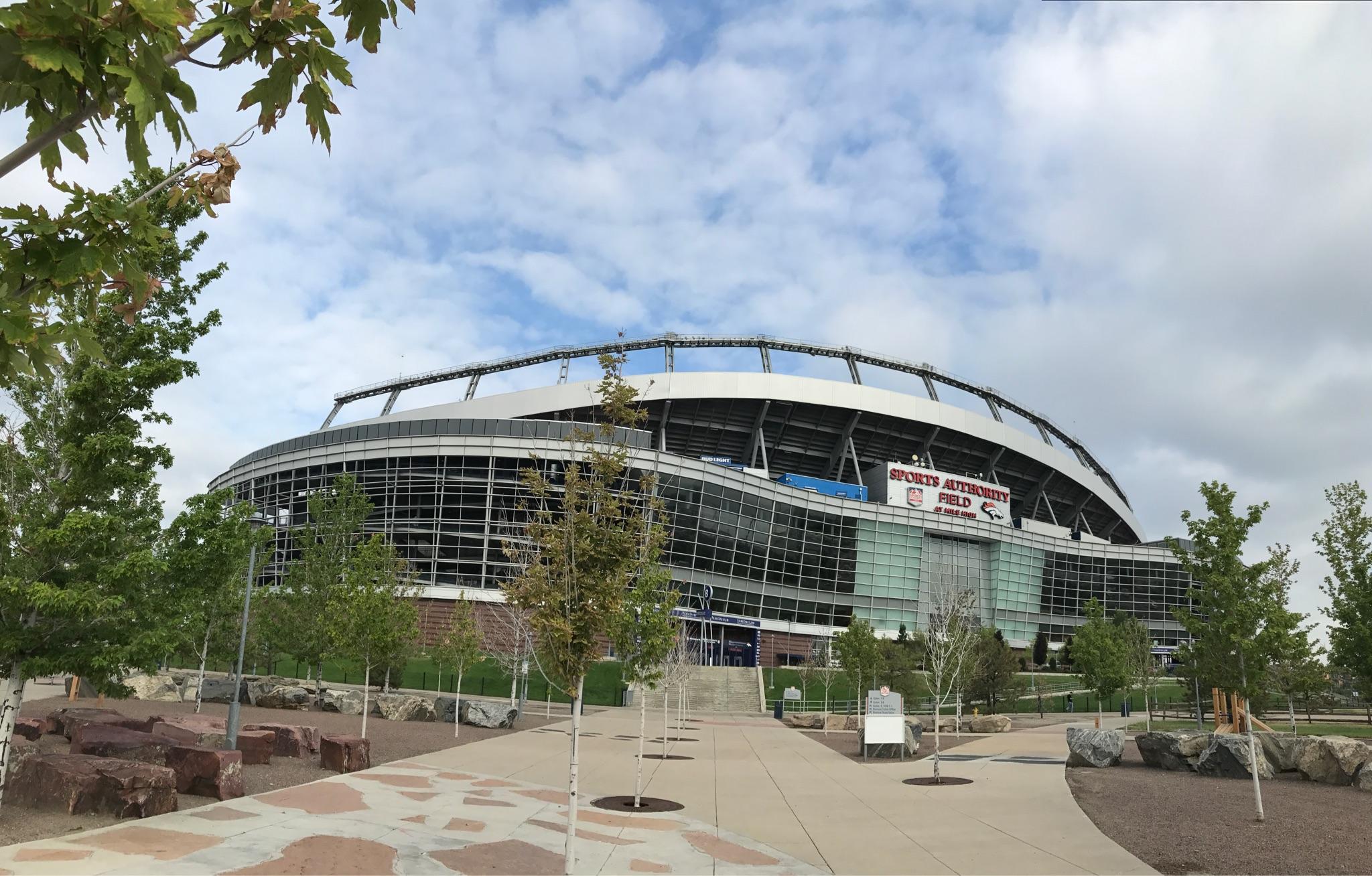 Large modern stadium building with trees and a paved walkway under a cloudy sky.