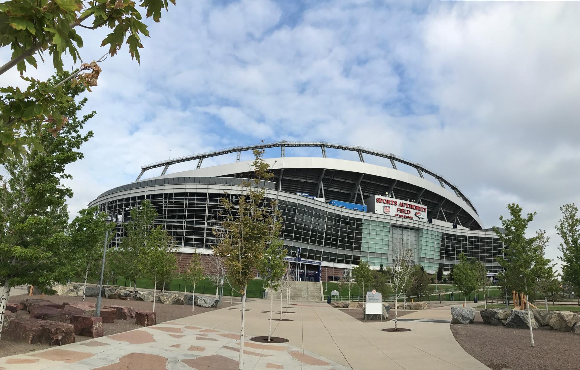 Large modern stadium building with trees and a paved walkway under a cloudy sky.