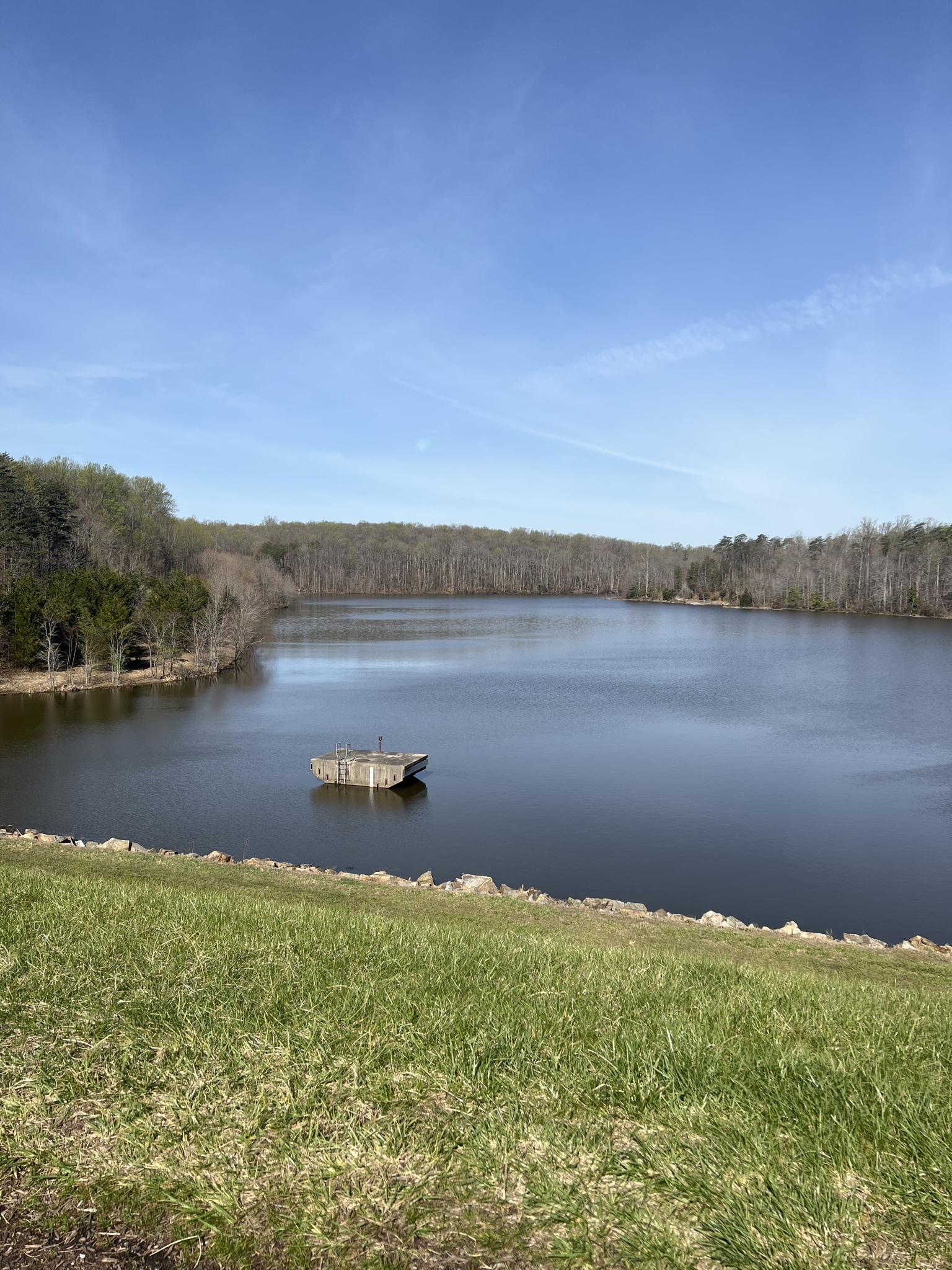 Lake with a small structure, surrounded by bare trees under a blue sky.