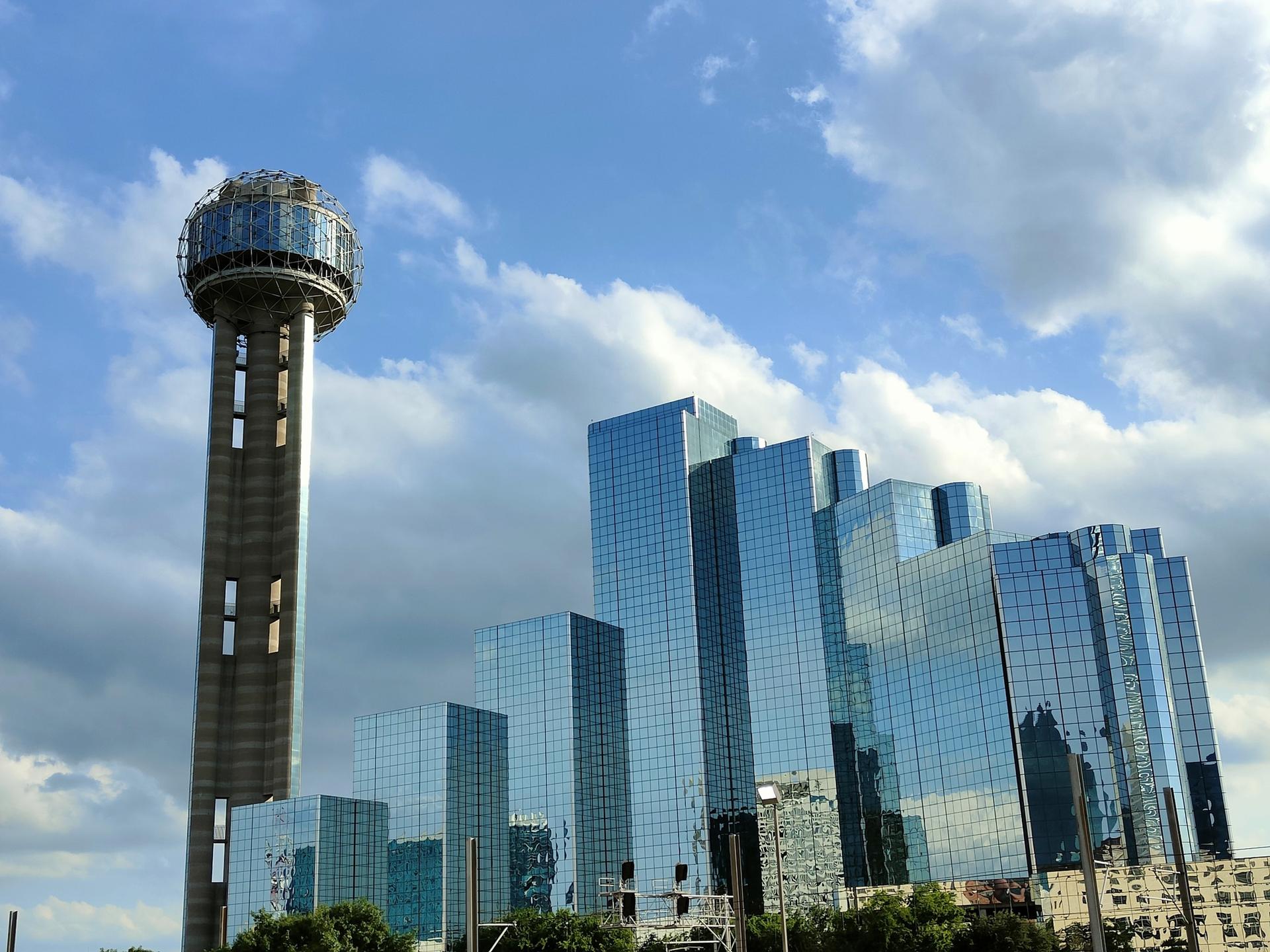 A tall, futuristic tower stands beside modern glass skyscrapers under a blue sky.