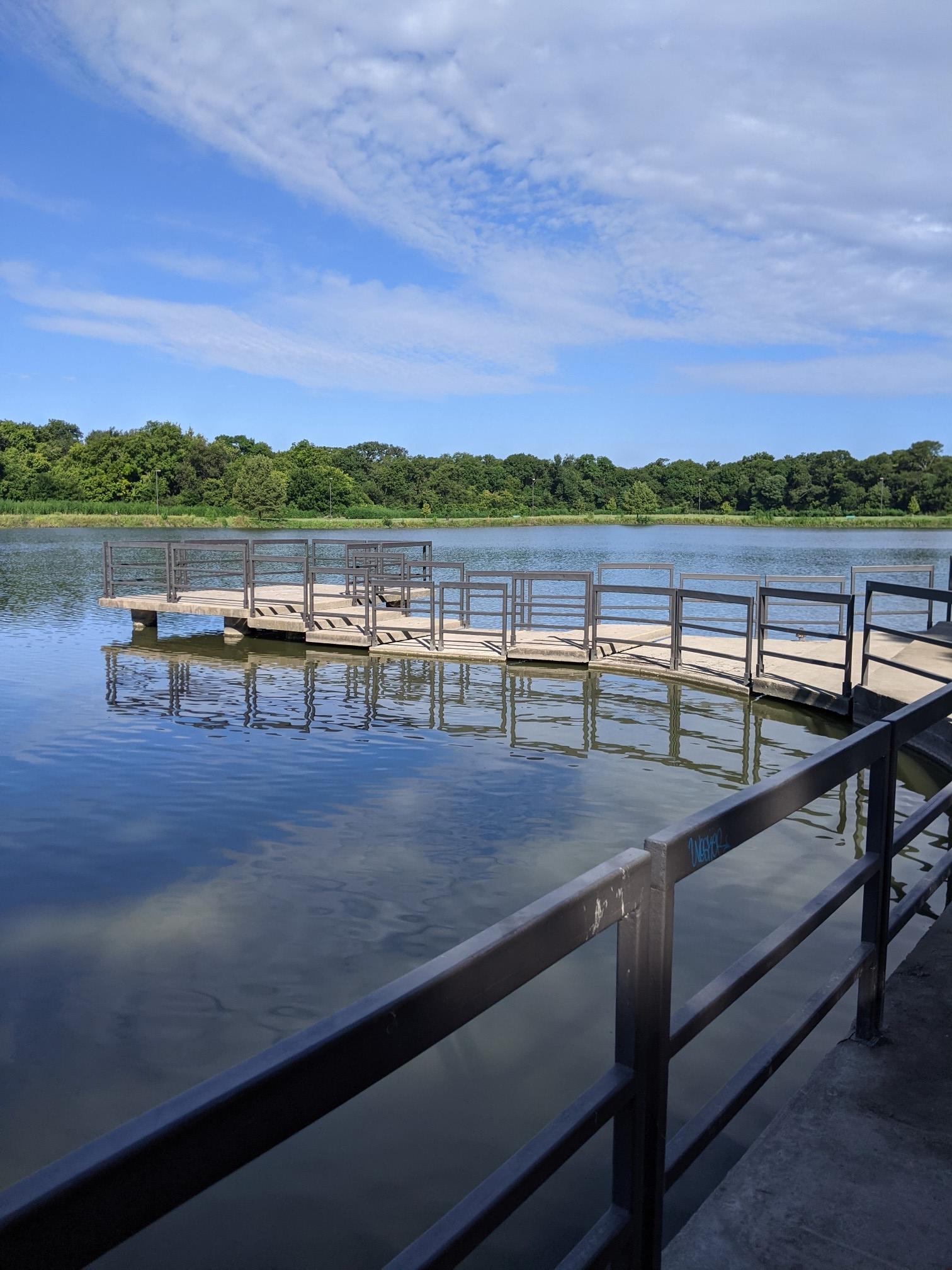 A winding wooden dock extends over a calm lake under a blue sky.