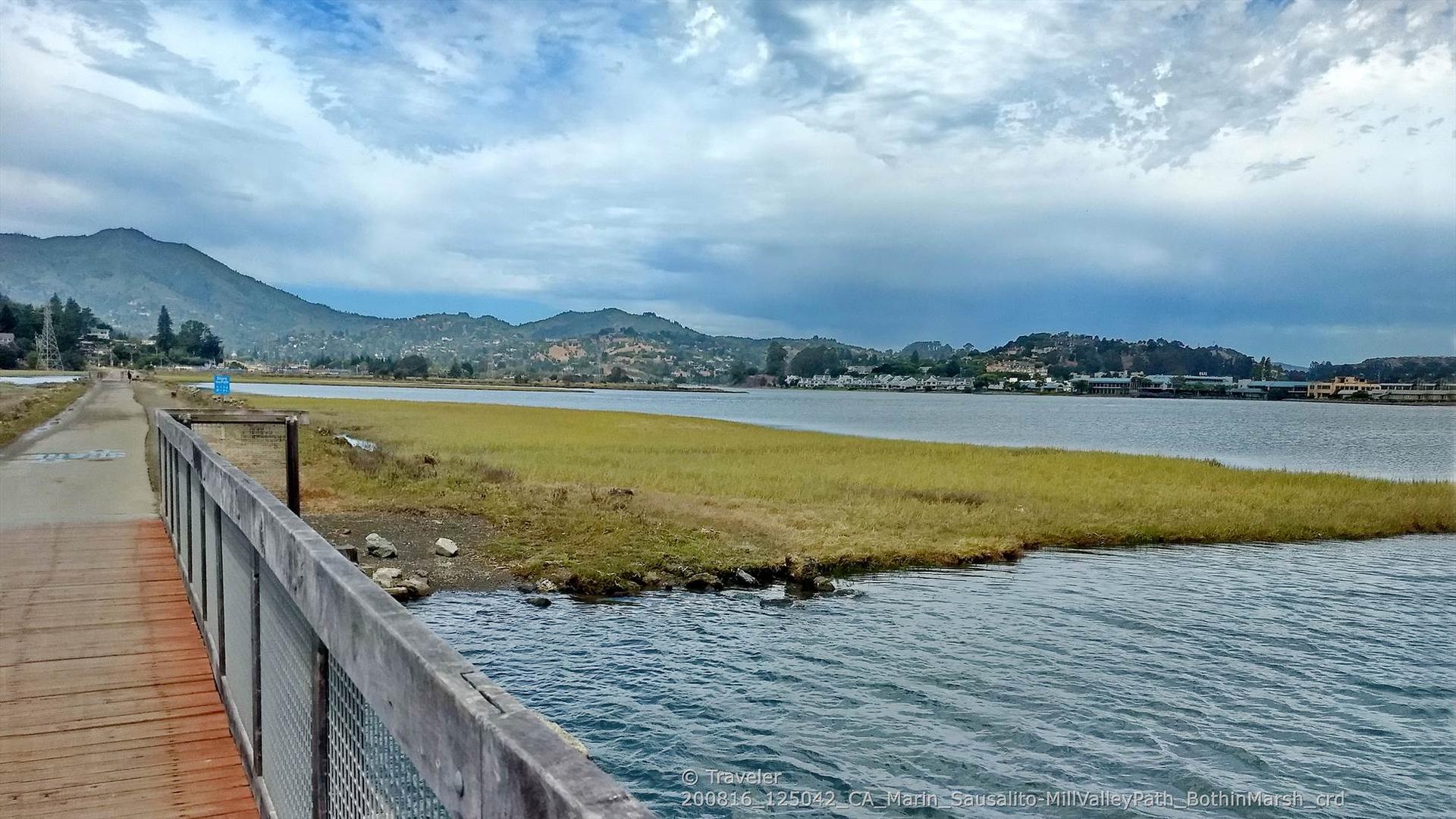 Wooden boardwalk overlooks a calm bay with distant mountains under an overcast sky.