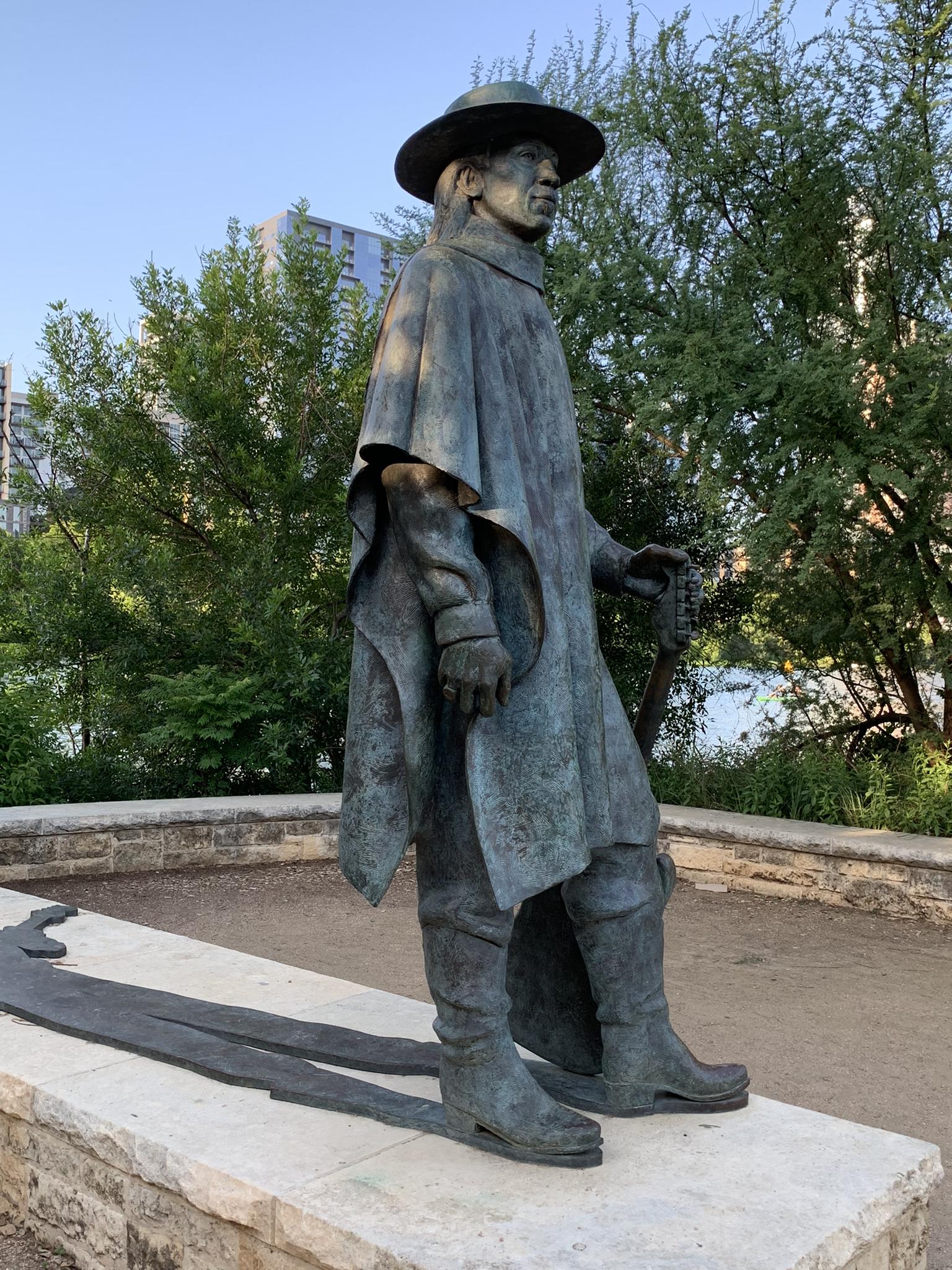 Bronze statue of a person in a hat and poncho, standing outdoors.