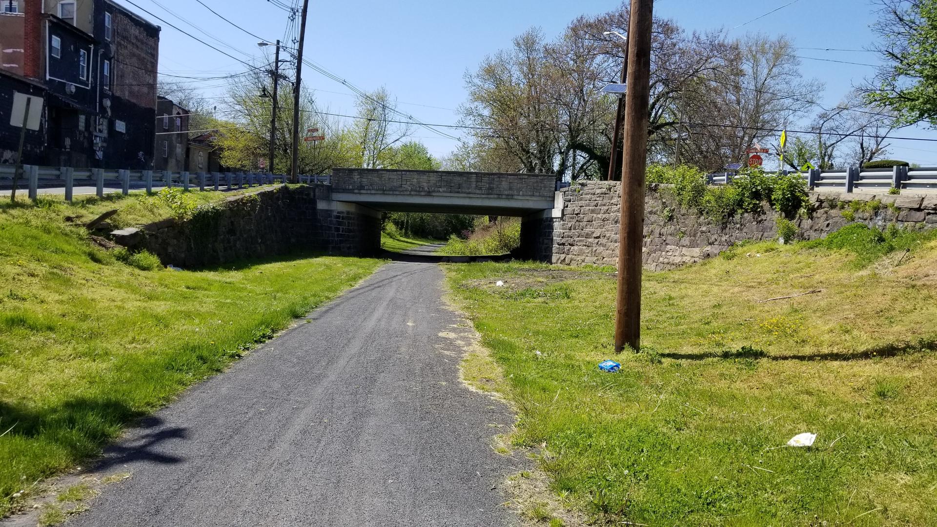 Paved path under a stone bridge with grassy banks on a sunny day.
