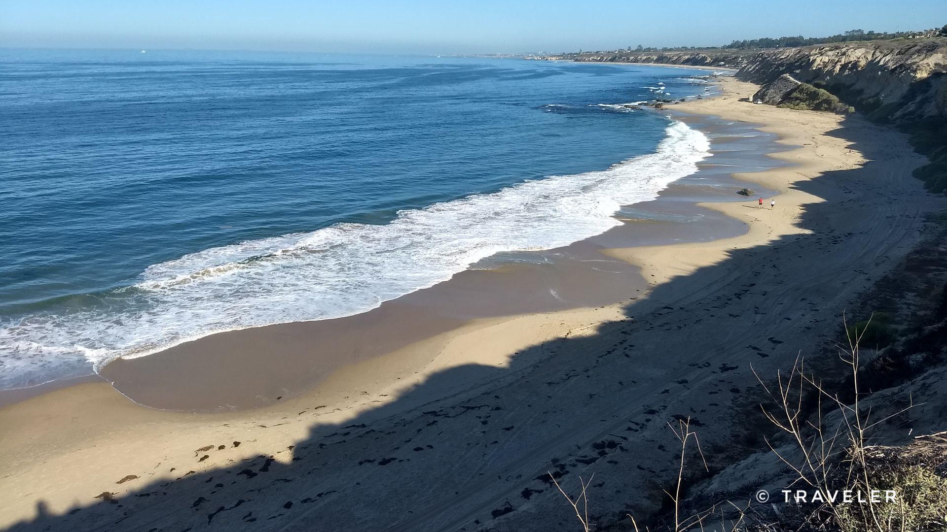 Sunny beach with crashing waves, coastal cliffs, and distant people.