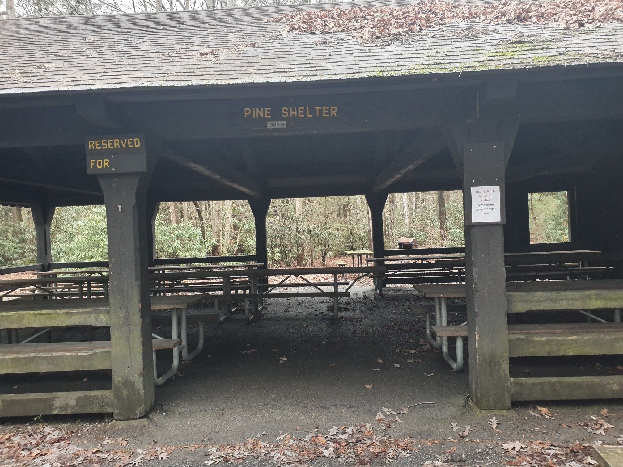 A large wooden picnic shelter with many tables in a forest. - Ramble Trail