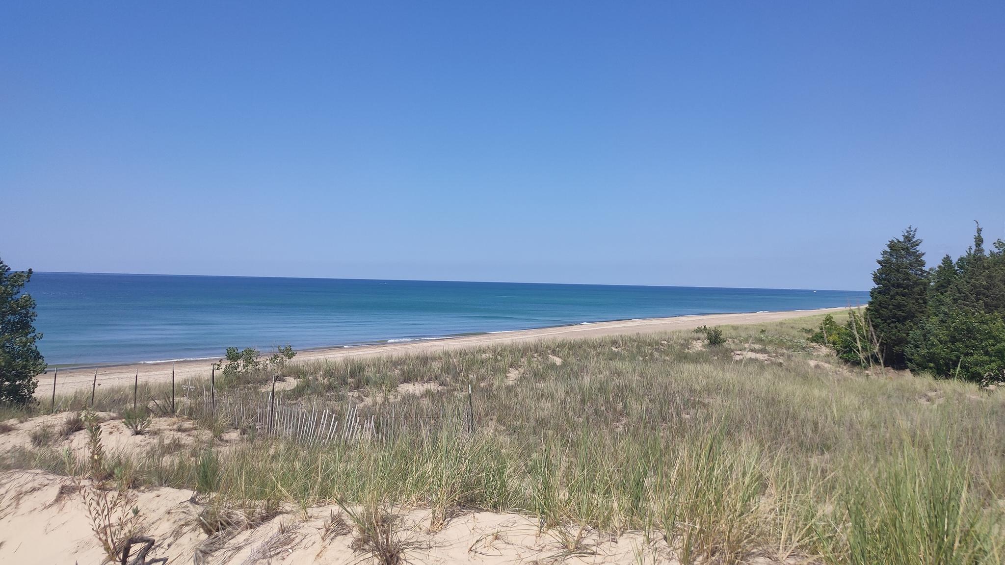 A wide sandy beach with grassy dunes stretches along calm blue water.