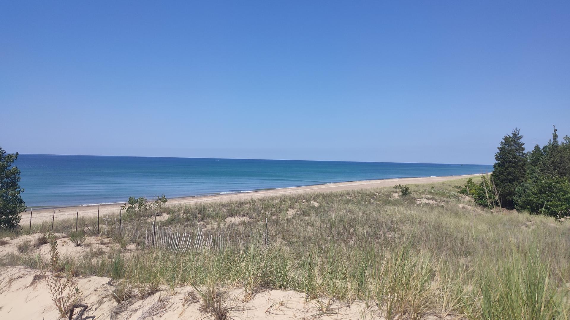 A wide sandy beach with grassy dunes stretches along calm blue water.