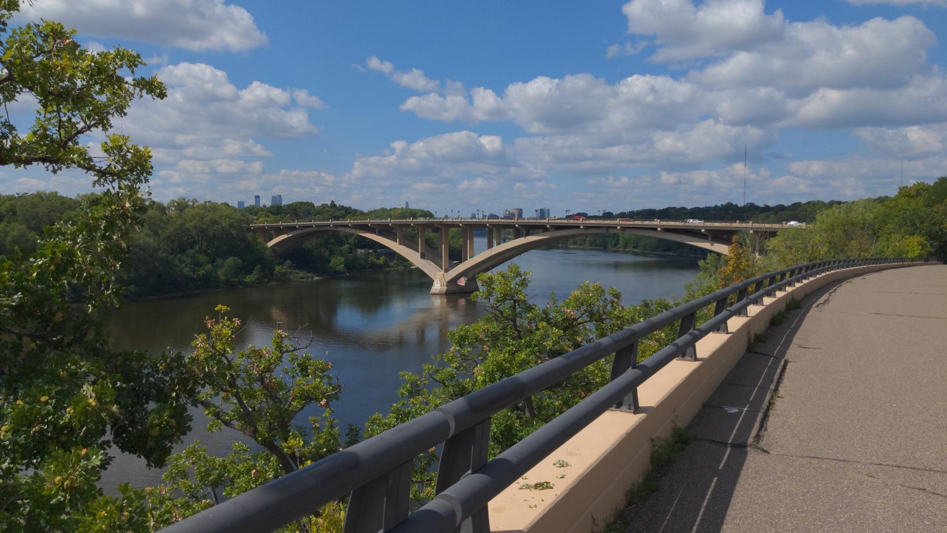 Arched bridge spans a wide river under a bright, cloudy sky.