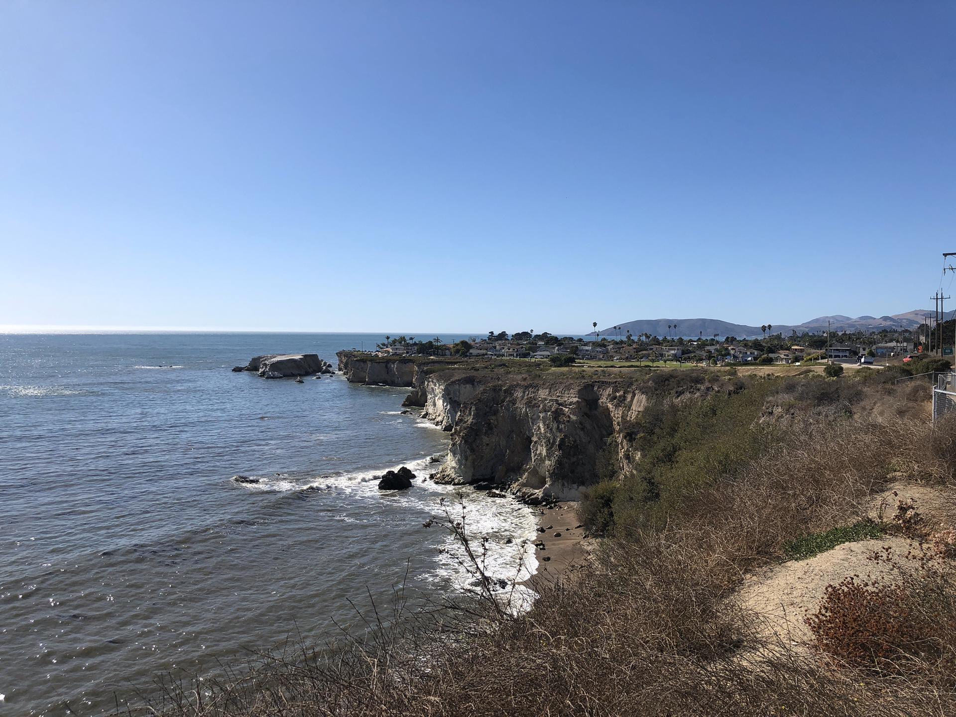 Ocean waves crash against rocky cliffs with a distant town under clear skies.