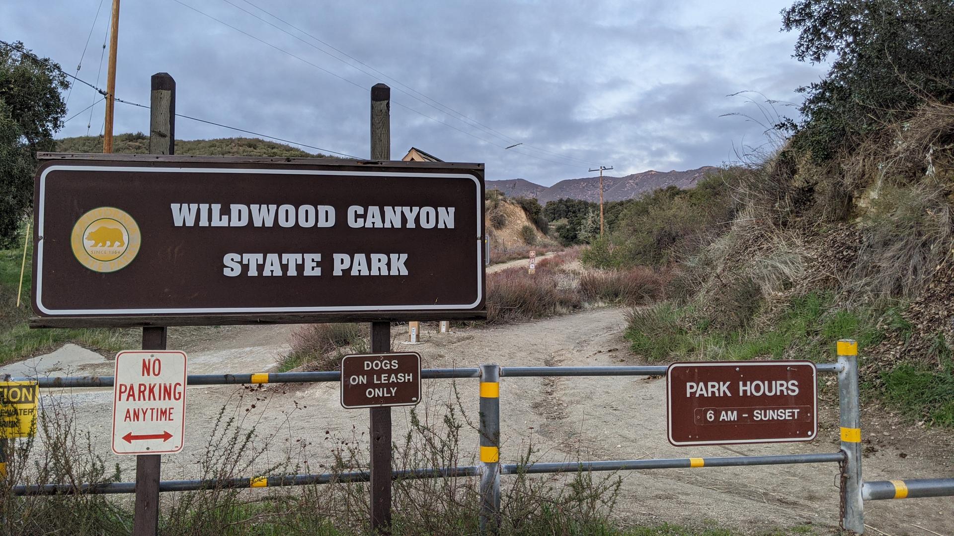 Park entrance sign and gate on a cloudy day, leading to a dirt path.