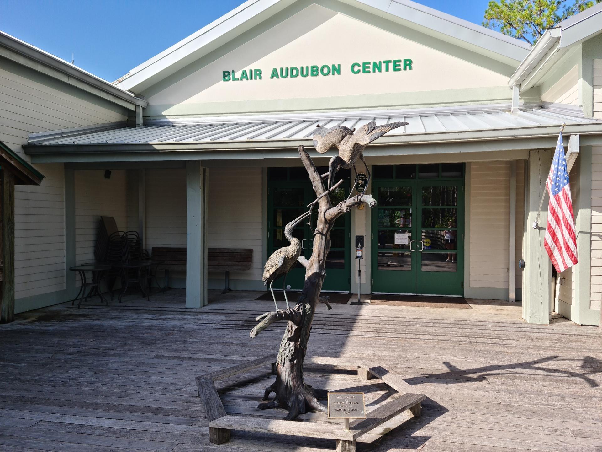 Building entrance with a bird sculpture on a wooden deck under bright sunlight.
