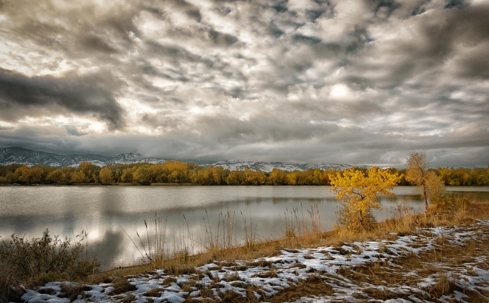 Serene lake reflecting a dramatic cloudy sky, with autumn trees and snow.