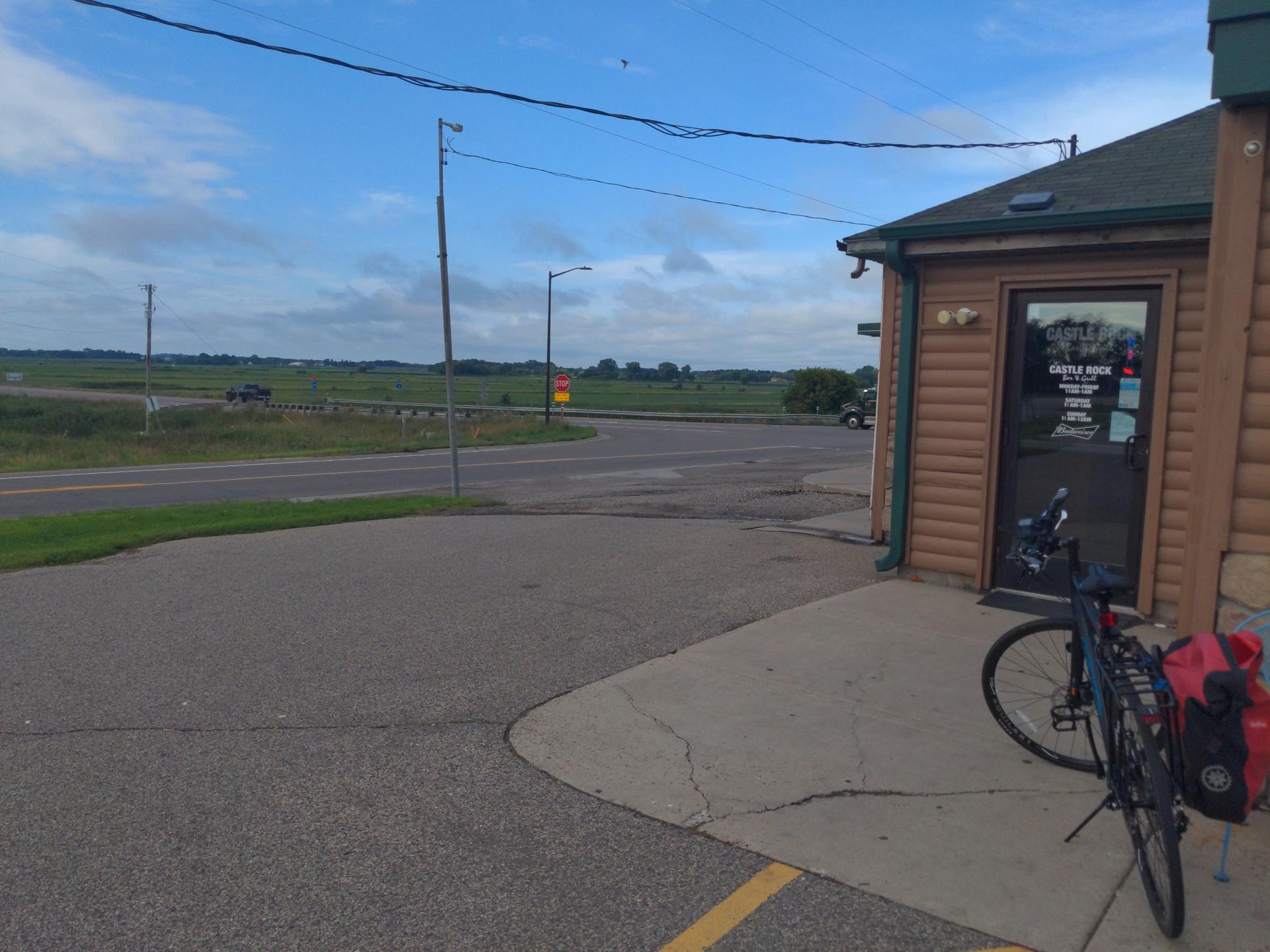 Bicycle with panniers parked beside a building near a road and open fields.