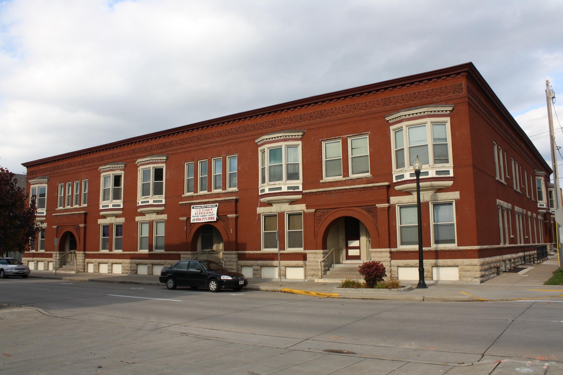 Large red brick building with many windows, parked cars on a street.