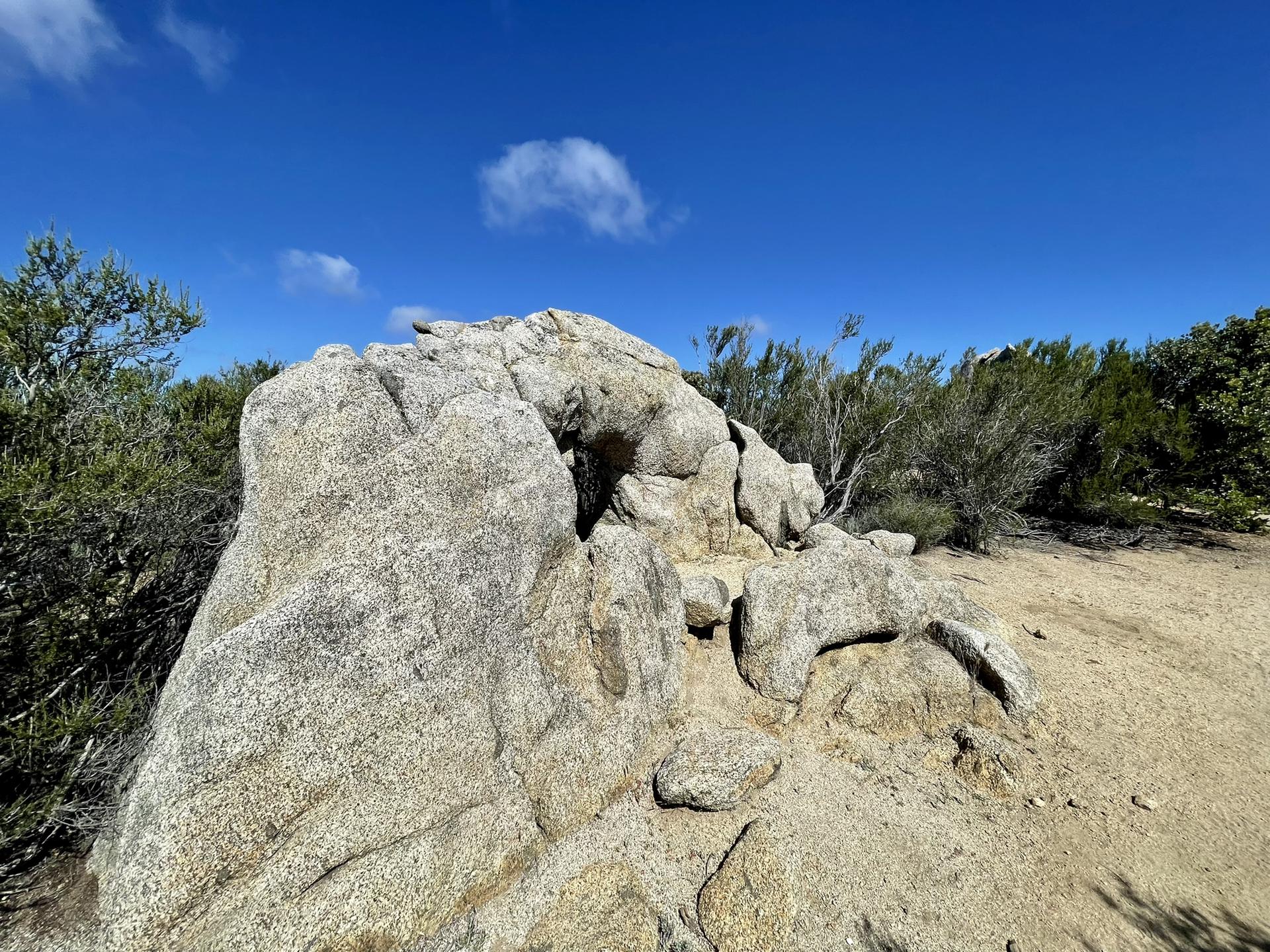 Large, textured rock formation on a dirt path with green bushes.