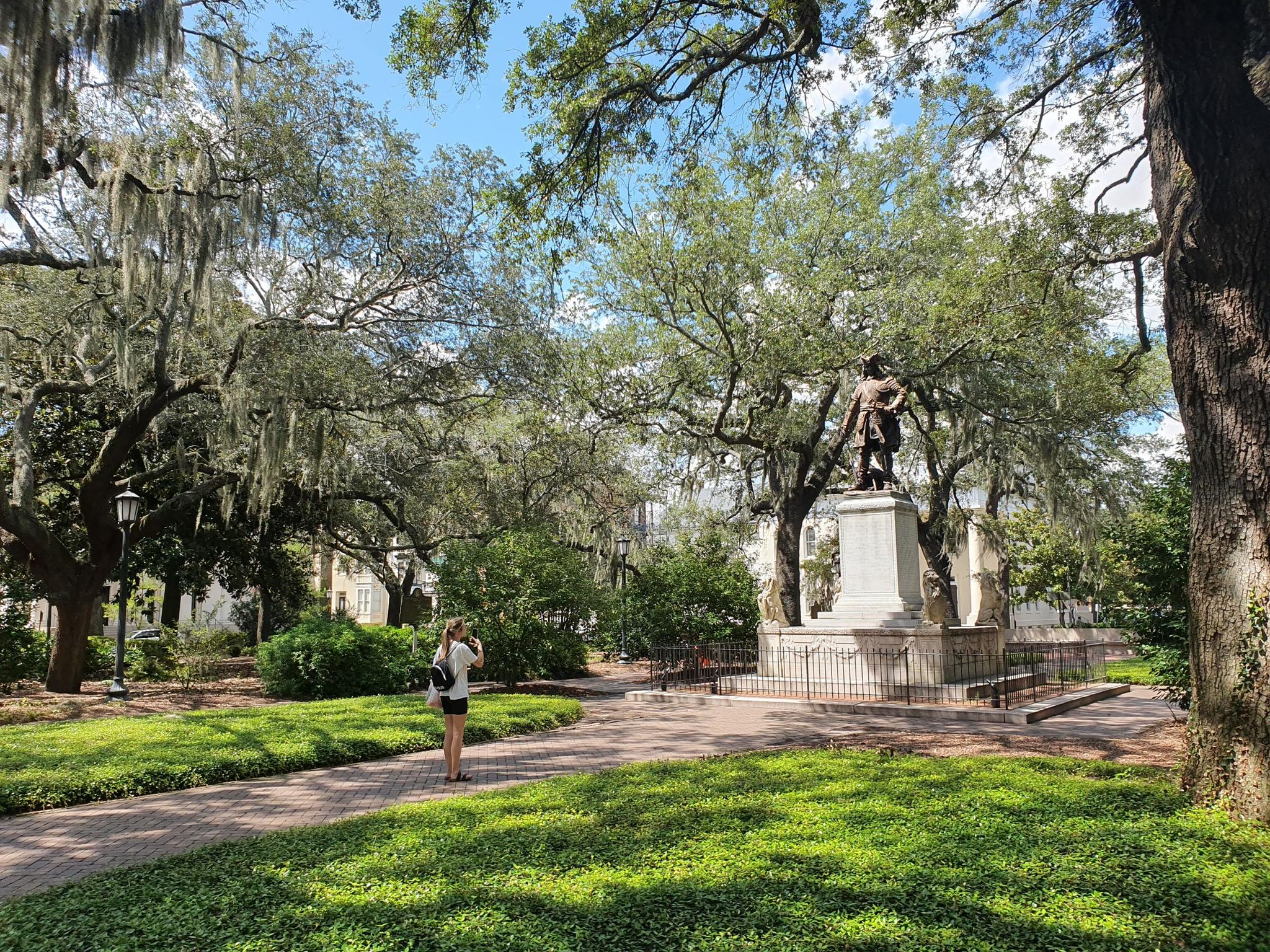 A woman stands on a path in a sunny park with a statue.