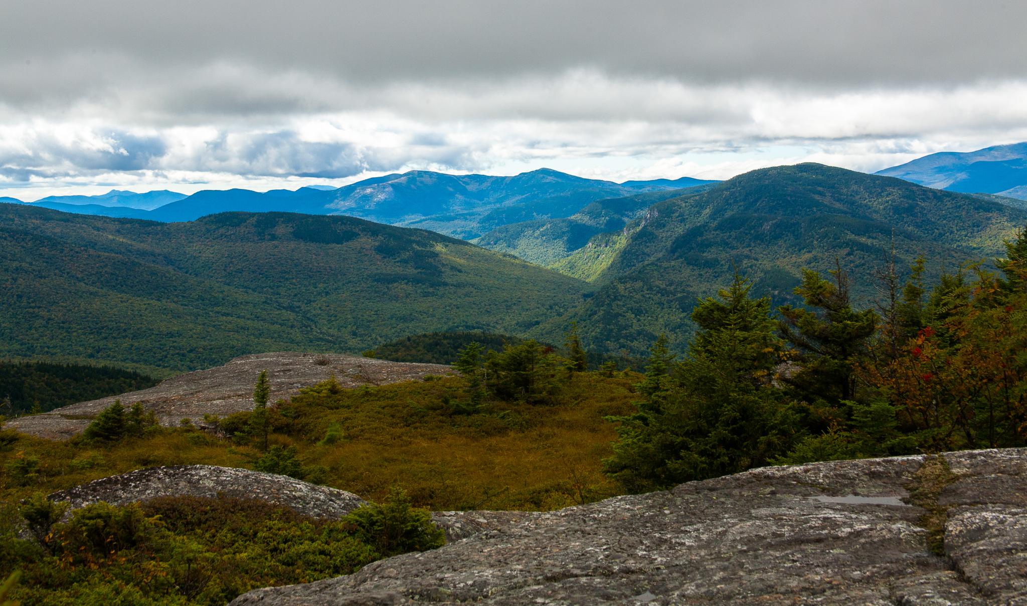 Dramatic mountain landscape with forested valleys and rocky foreground under cloudy sky.
