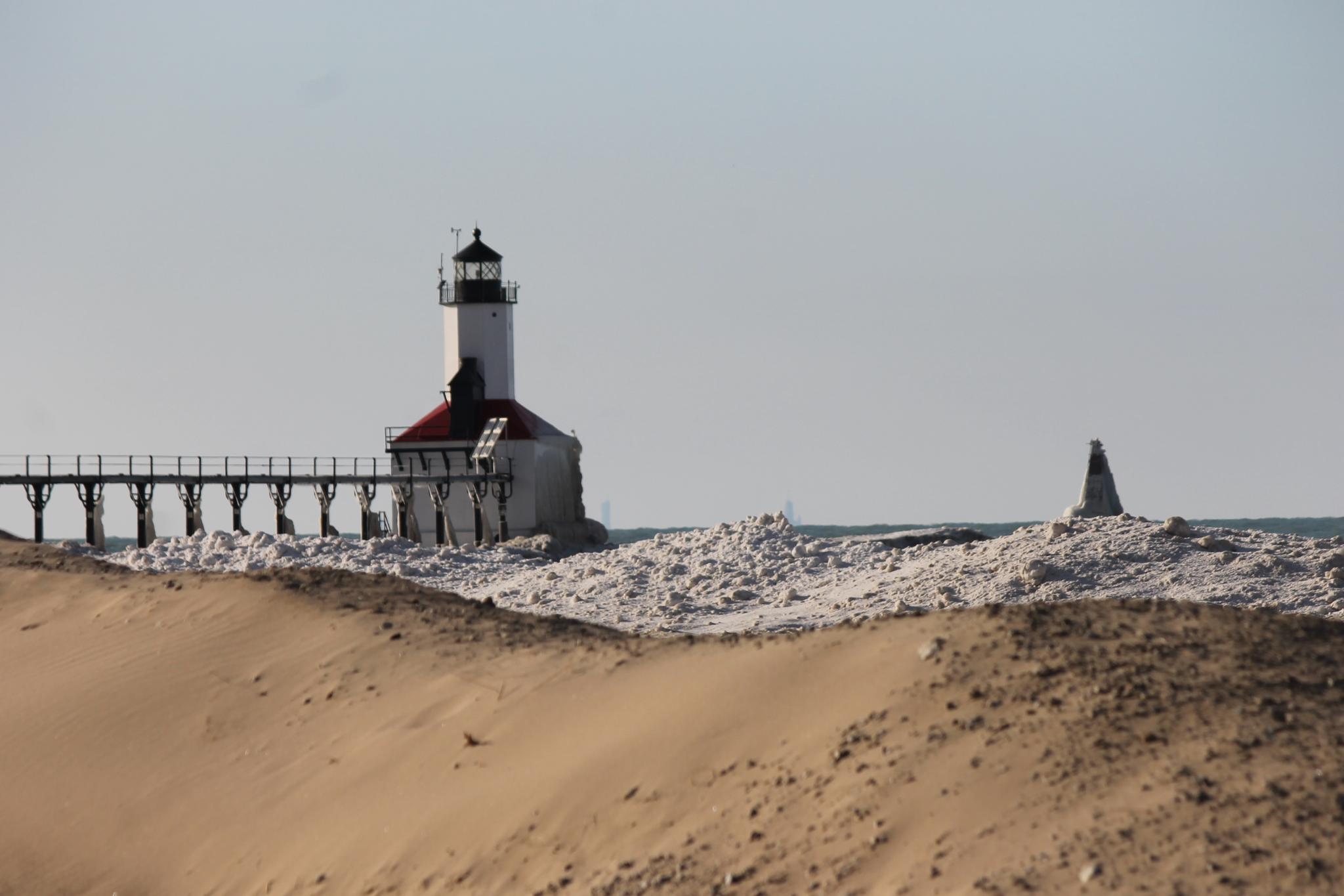 Lighthouse and pier over icy water with sandy dunes in bright daylight.