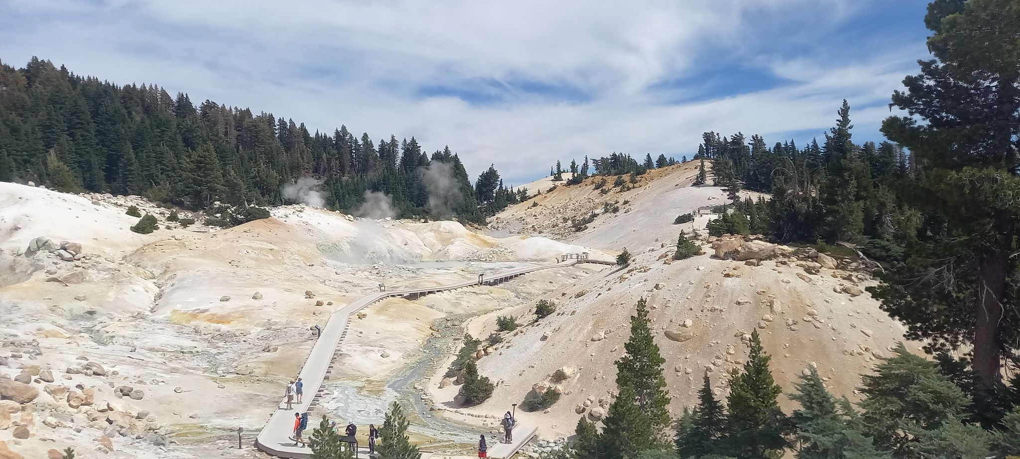 Geothermal area with steaming vents, light-colored ground, and people on boardwalks. - Bumpass Hell Trail