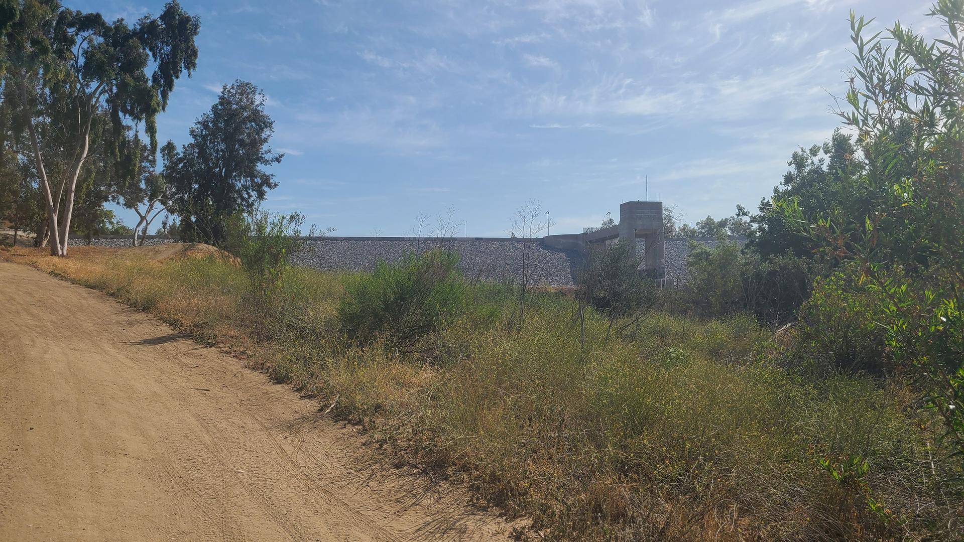 A dirt path leads towards a large concrete dam under a bright sky.
